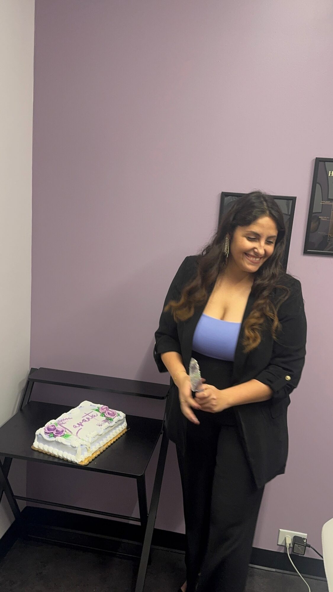 Woman with long hair smiling, standing next to a table with a cake, purple wall background.