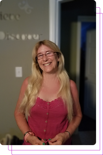 Smiling woman with long blonde hair and glasses, wearing a pink sleeveless top, standing indoors.