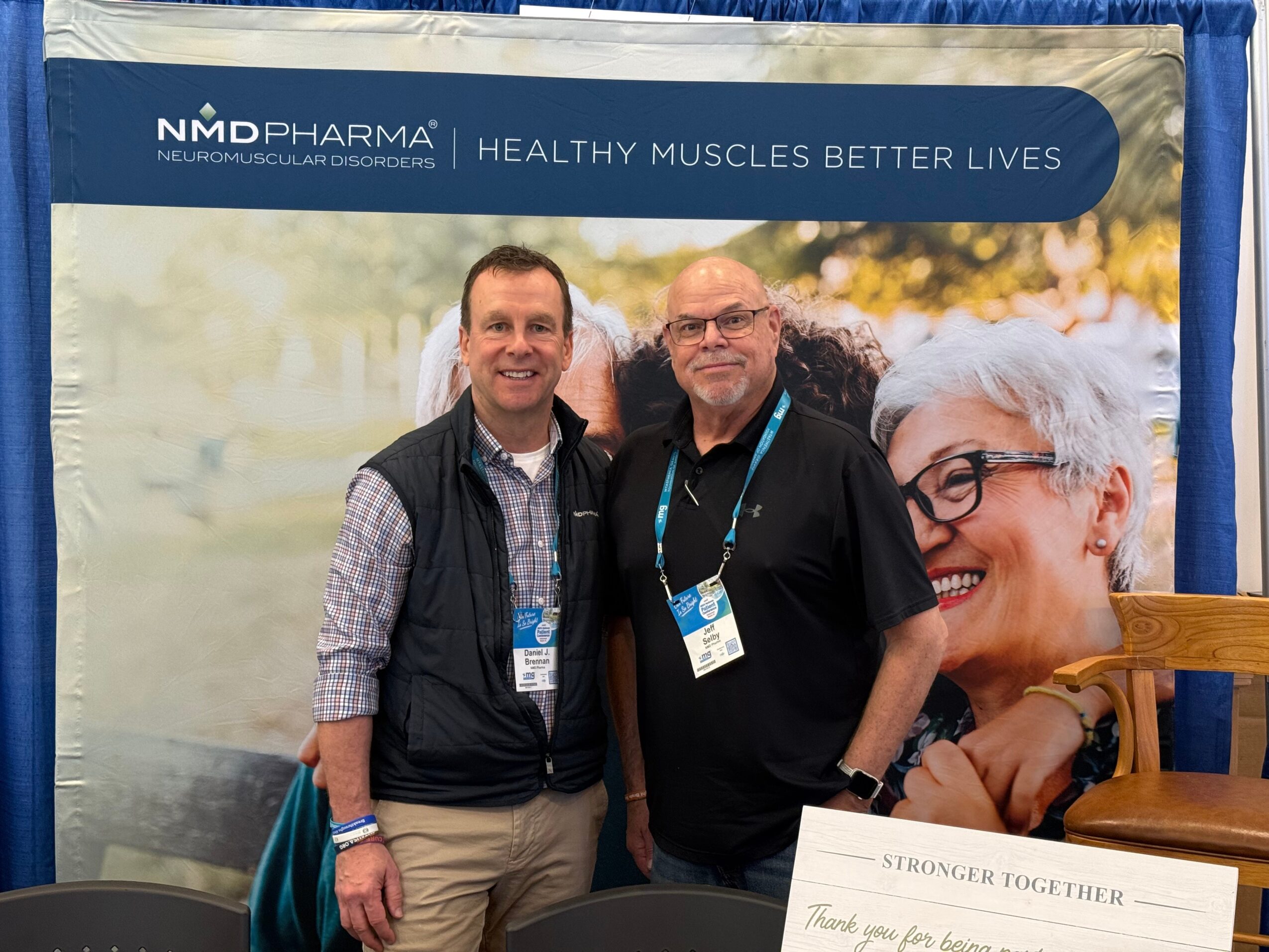 Two men standing in front of a promotional banner, smiling, with a large image of an elderly woman in the background.