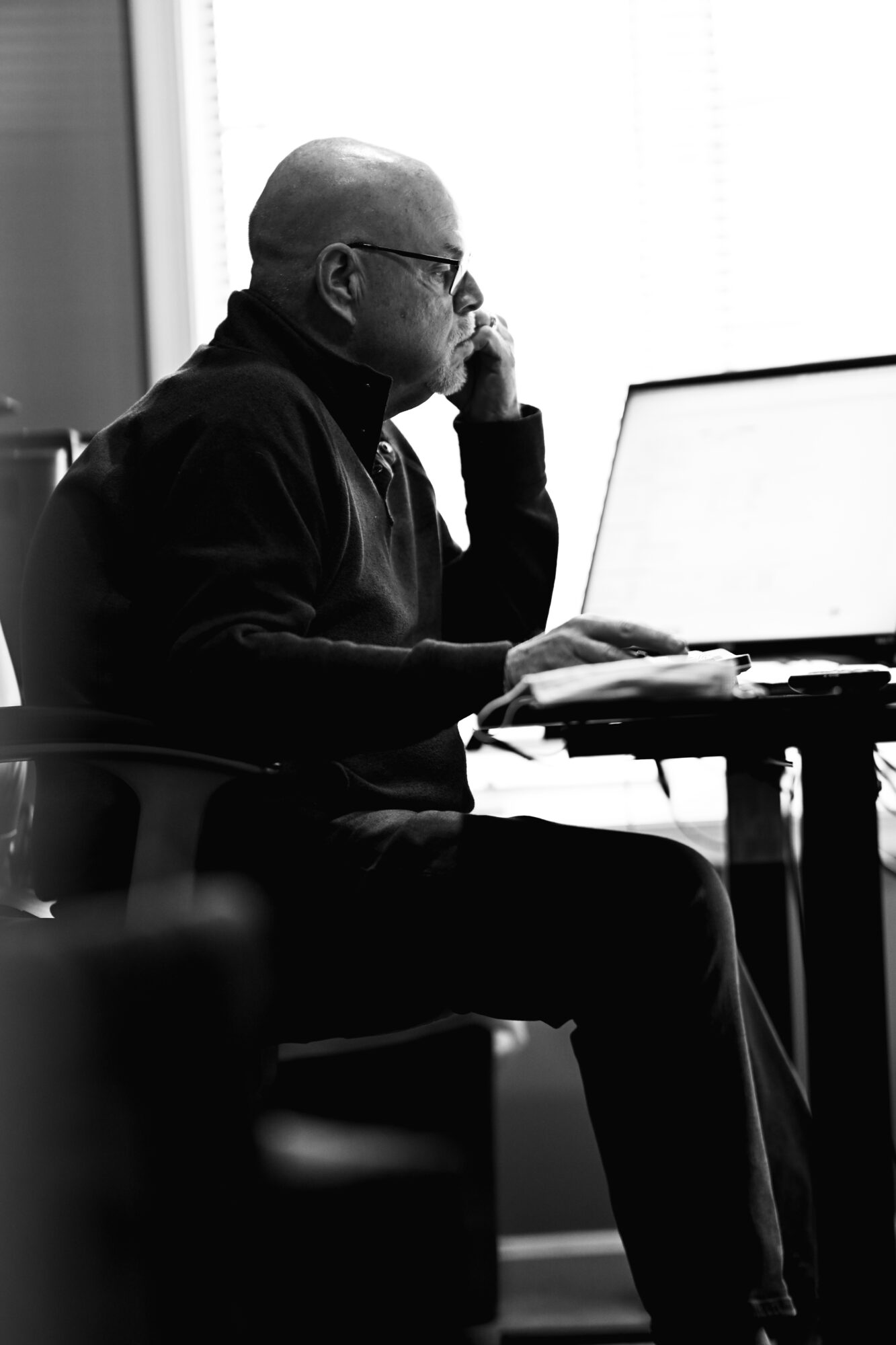Man with glasses working on a laptop at a desk, sitting in a chair near a window, in black and white.