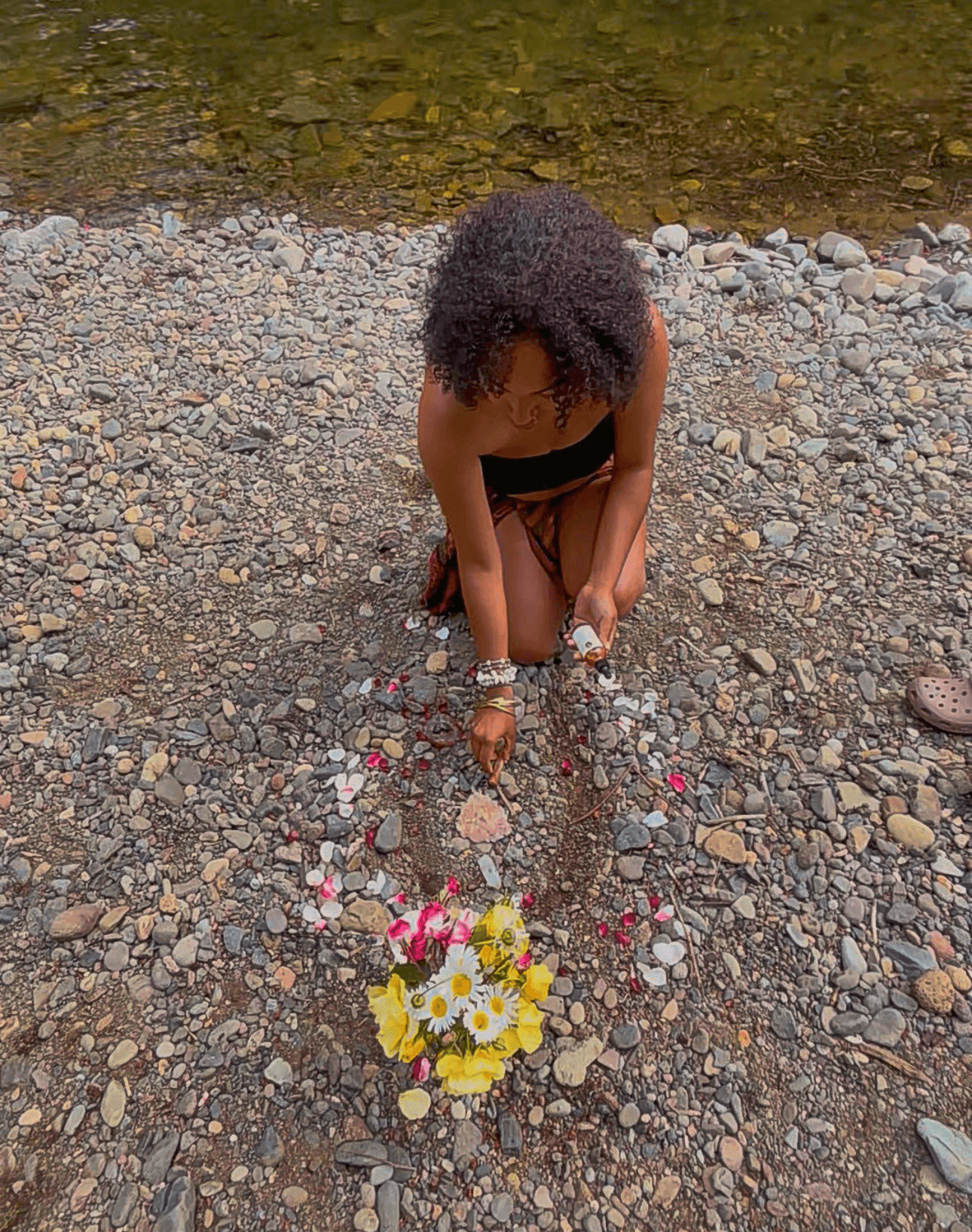 Child with curly hair bends down on rocky ground near water, placing flowers in a small arrangement.