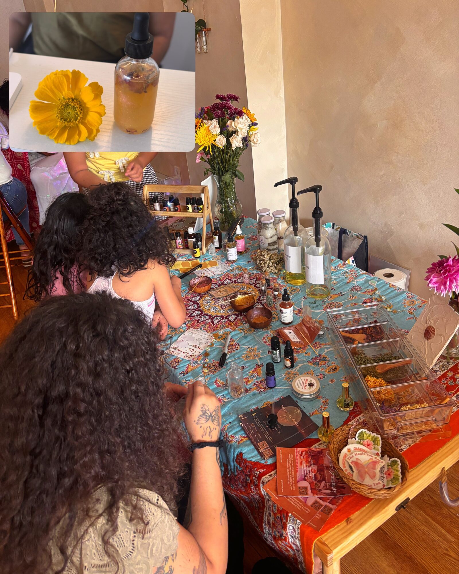 People gathered around a table with various bottles, flowers, and decorative items, in a cozy indoor setting.