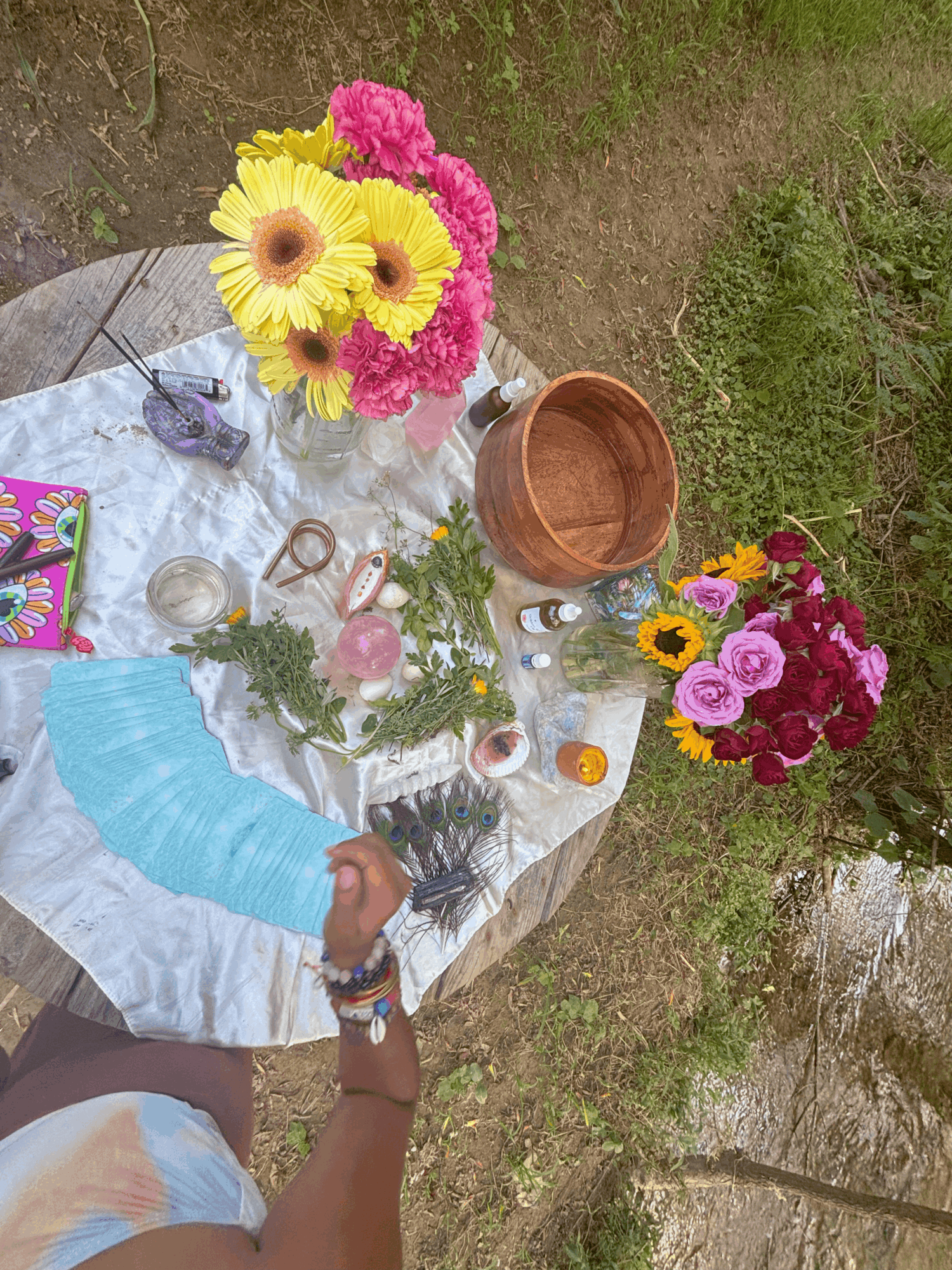 Outdoor table with colorful flowers, jewelry, and decorative items, set on grass with a person reaching for an object.
