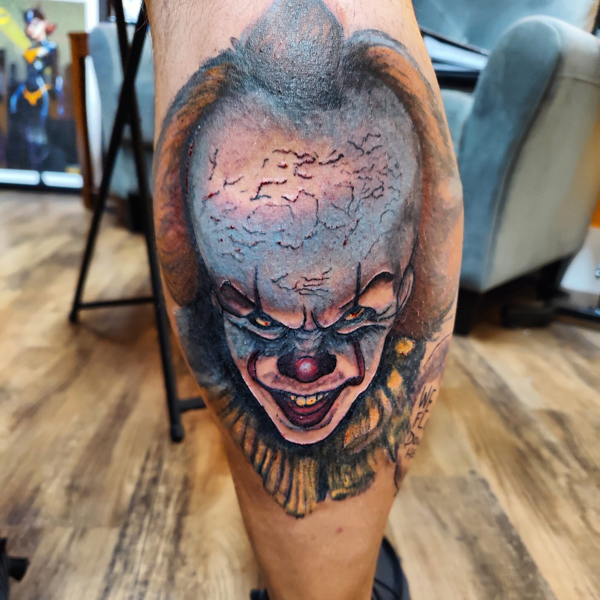 Tattoo of a clown with a sinister smile, bald head, and dark makeup around eyes, on a person's leg.
