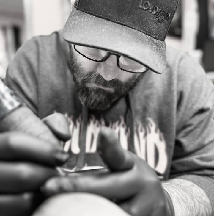 Man with glasses and a beard wearing a cap and hoodie, writing or drawing on a surface, in black and white.