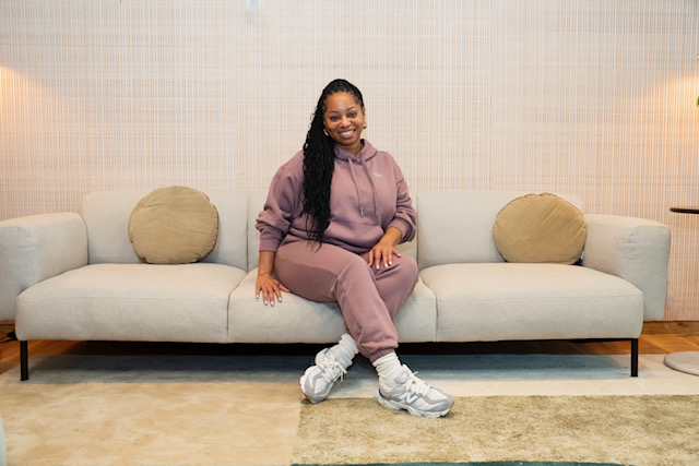 Woman sitting on a light-colored sofa with two beige pillows, smiling, in a room with beige walls and a carpeted floor.