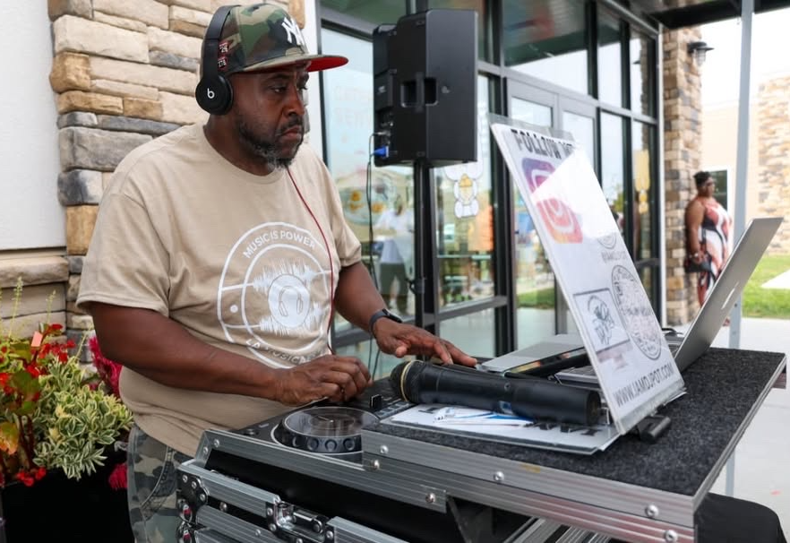 DJ wearing headphones and a cap, operating equipment outdoors near a building with large windows and stone walls.