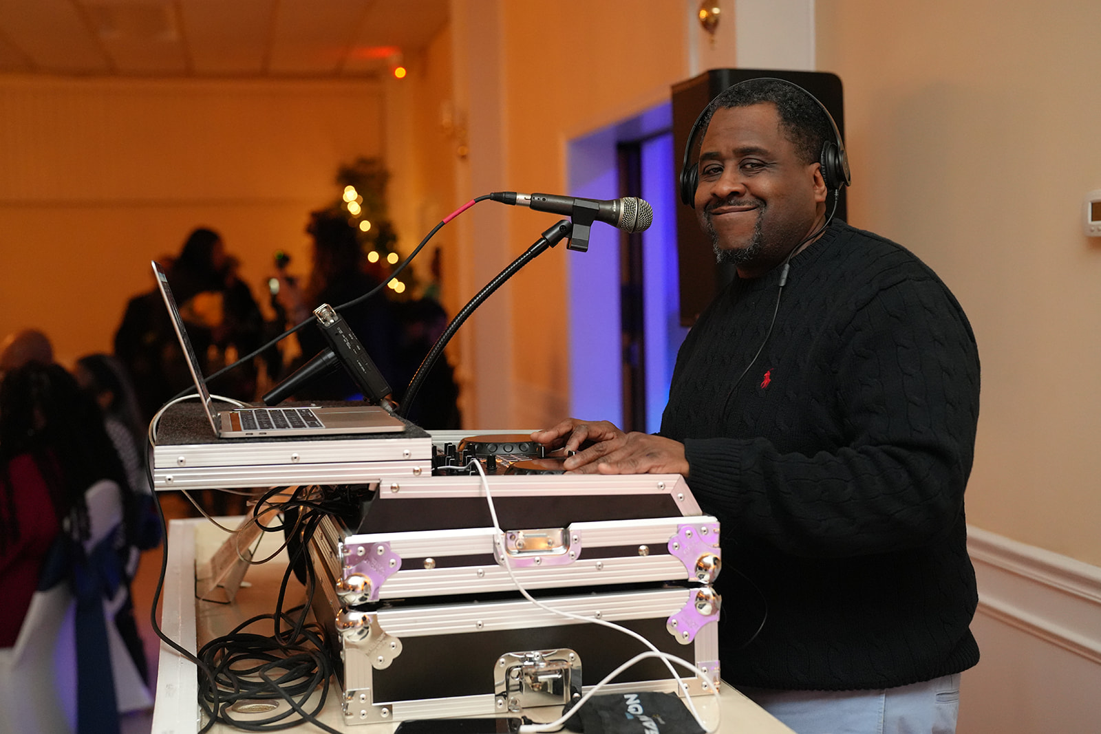 DJ smiling at turntables and laptop during event, with people in background, warm lighting, and decorated room.