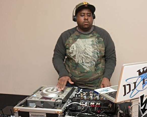 DJ wearing headphones and a camouflage shirt, standing behind DJ equipment, with a neutral background.