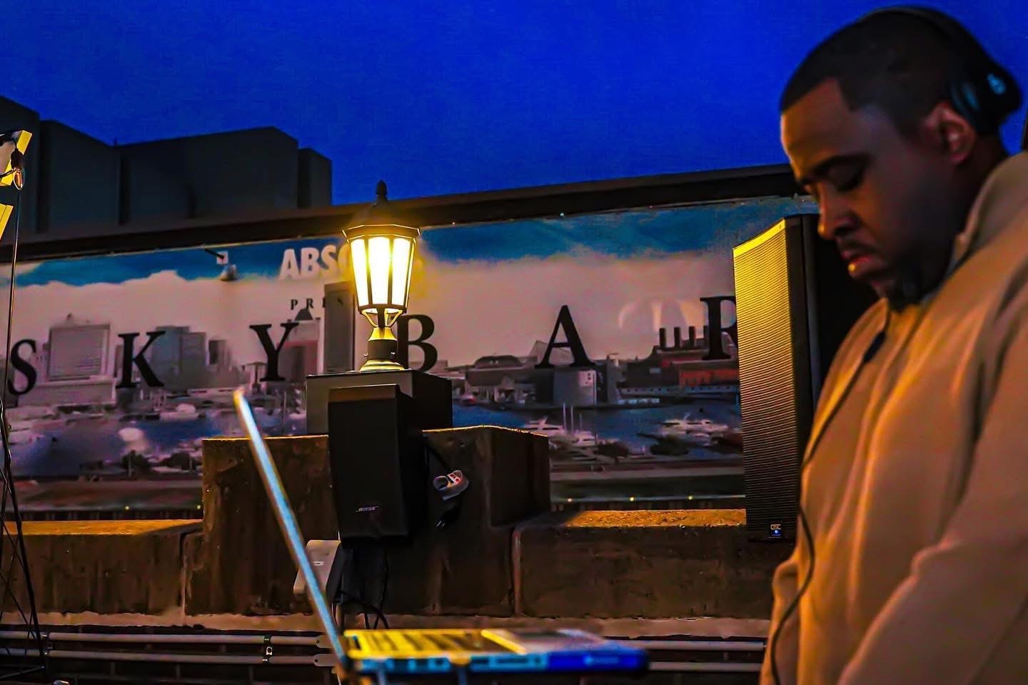 Person with dark hair in a beige jacket stands outdoors at night near a railing with a cityscape background.