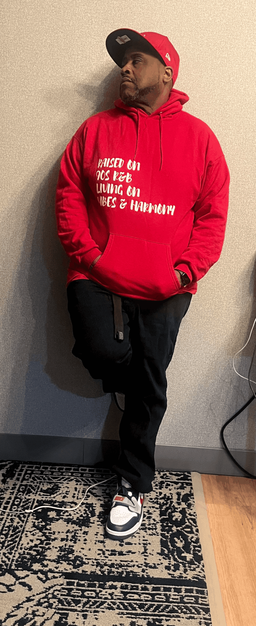 Person standing against a wall wearing a red hoodie, black pants, and sneakers, with hands in pockets.