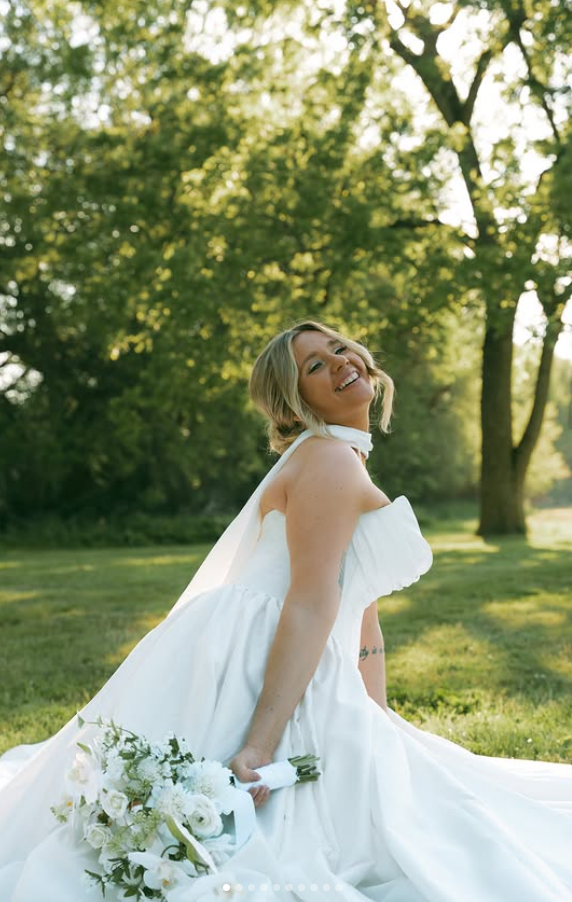 Smiling woman in a white dress sitting outdoors on grass with trees in background.