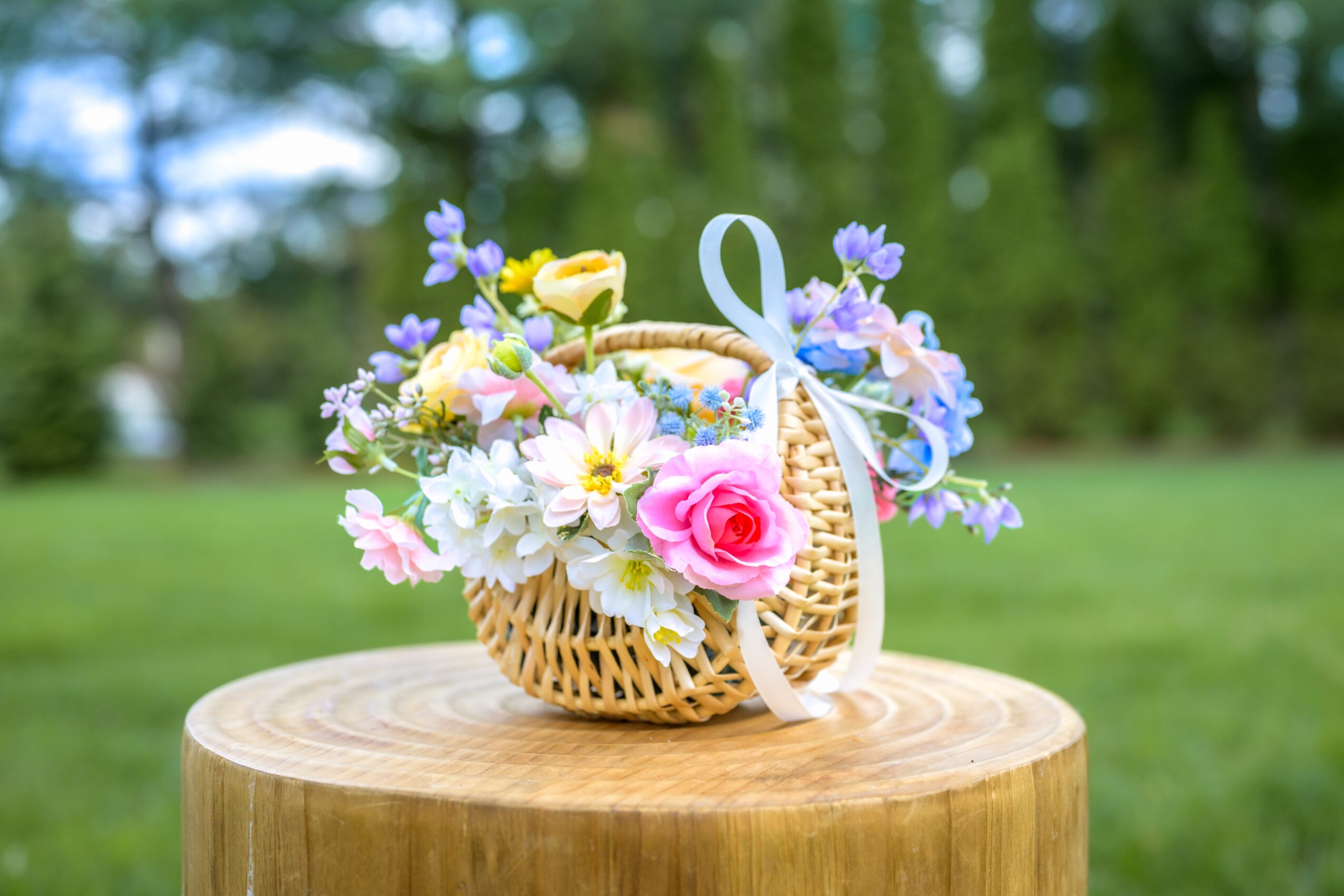Basket filled with colorful flowers and a small yellow bird, placed on a wooden surface outdoors.