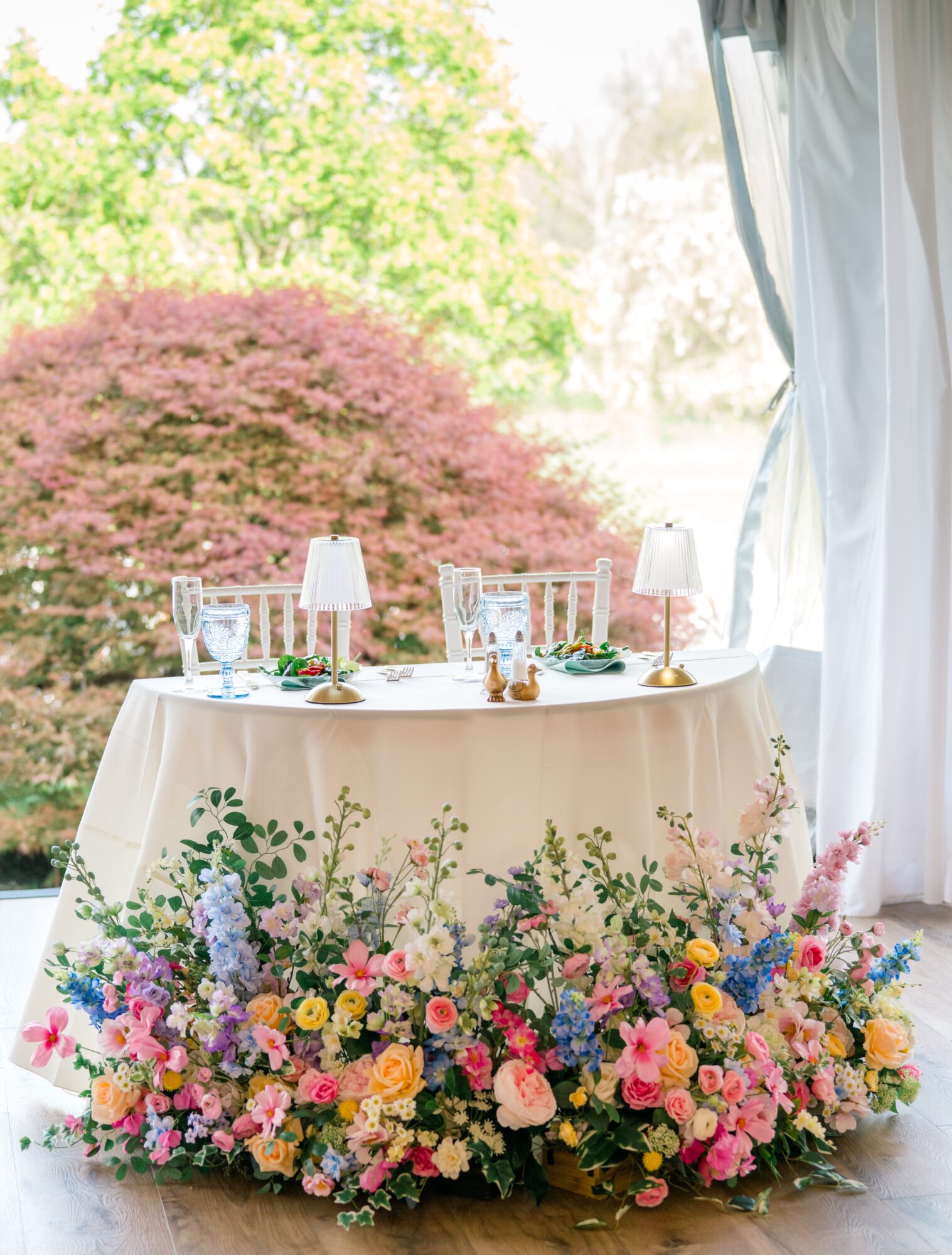 Table with floral arrangement, two lamps, and glassware outdoors, with trees and pink bush in background.