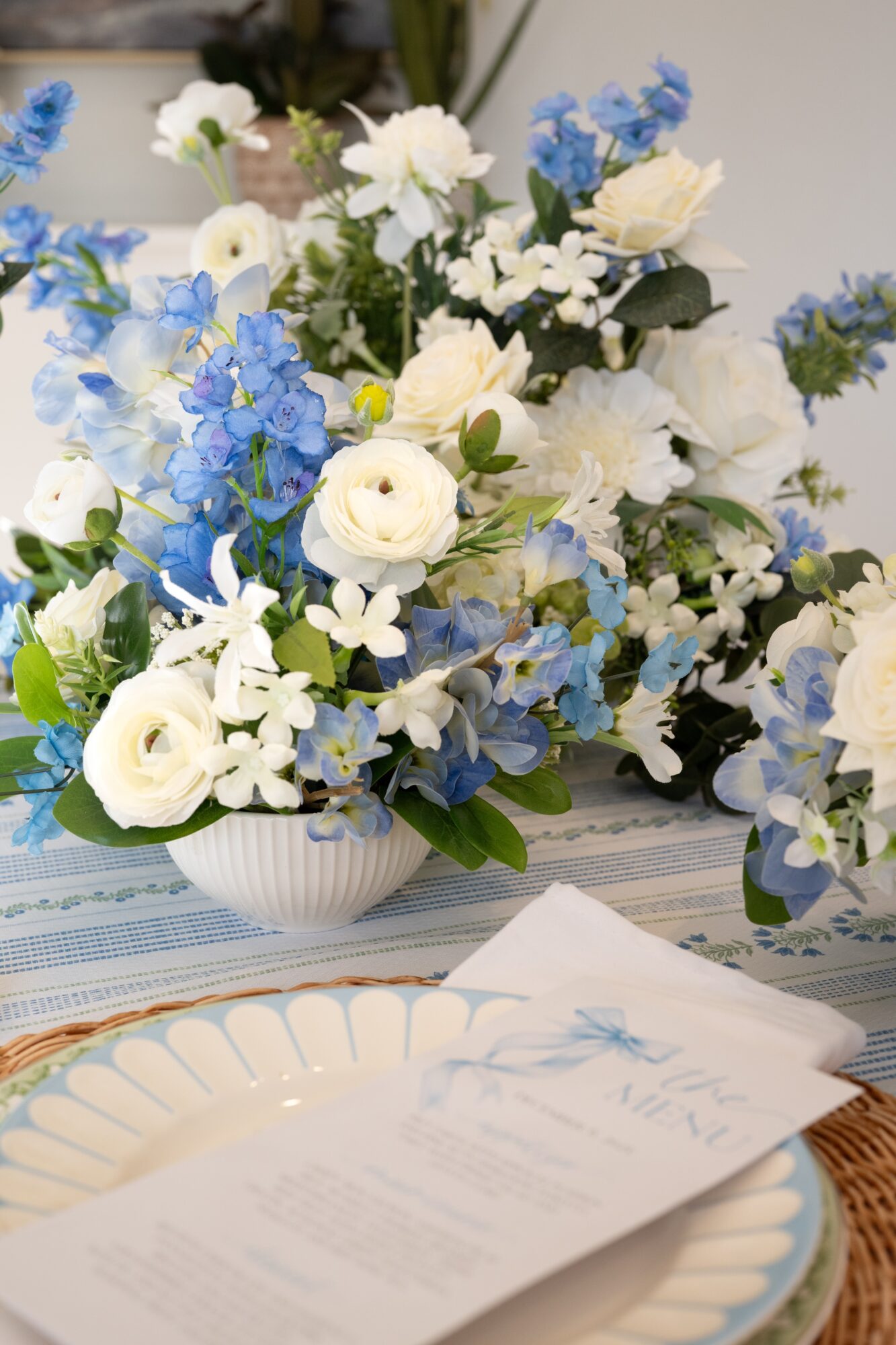 Flower arrangement with white and blue flowers in a white vase on a table, with a plate and napkin nearby.