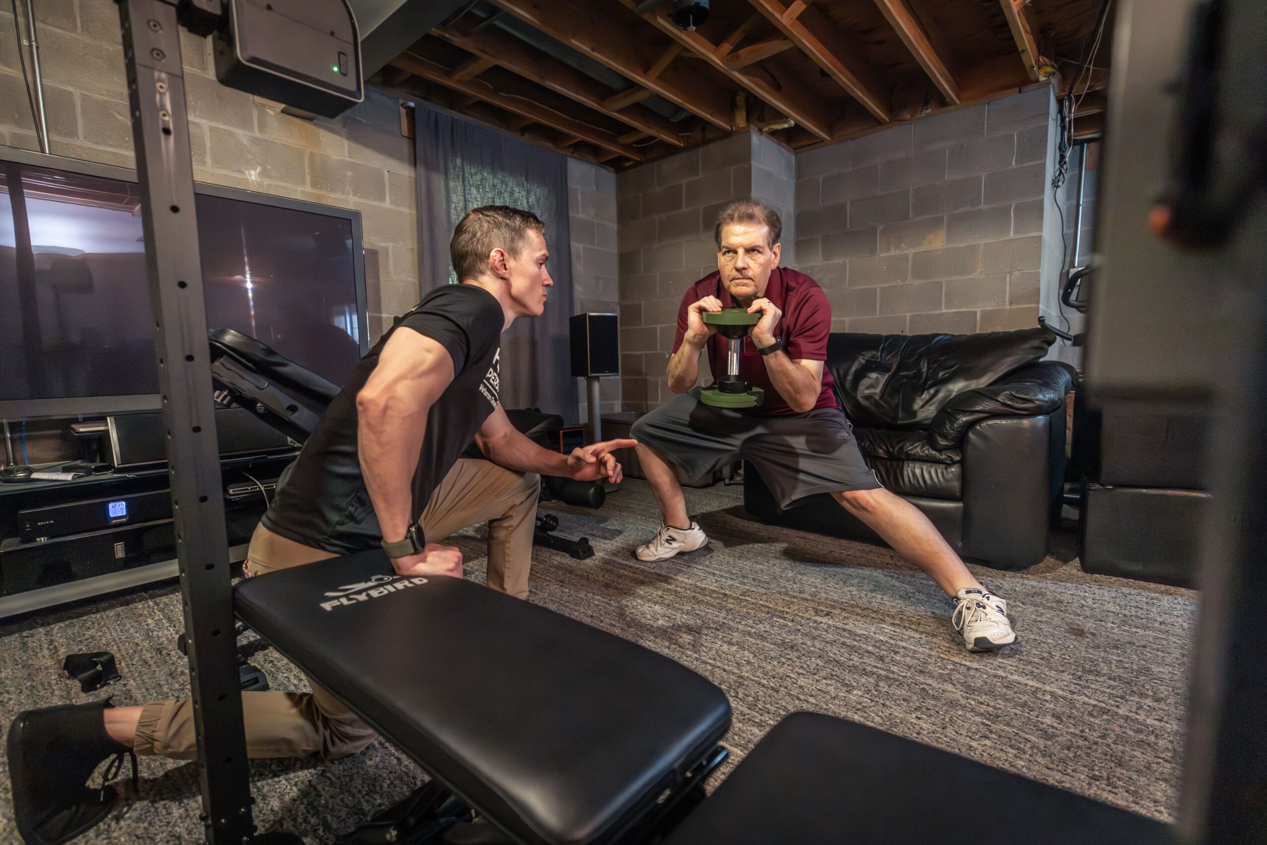 Two men are exercising in a basement with concrete walls and a wooden ceiling, one holding a weight and the other in a stretching pose.