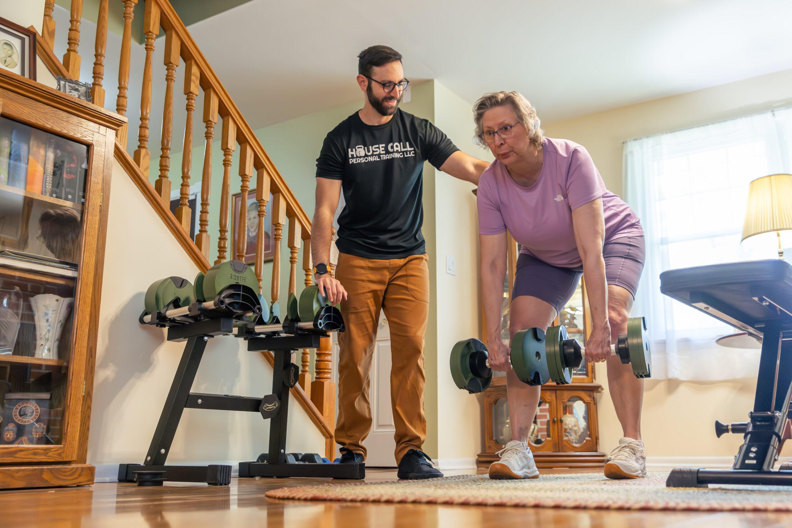 A man assists an elderly woman with dumbbells in a living room, with a staircase and furniture in the background.