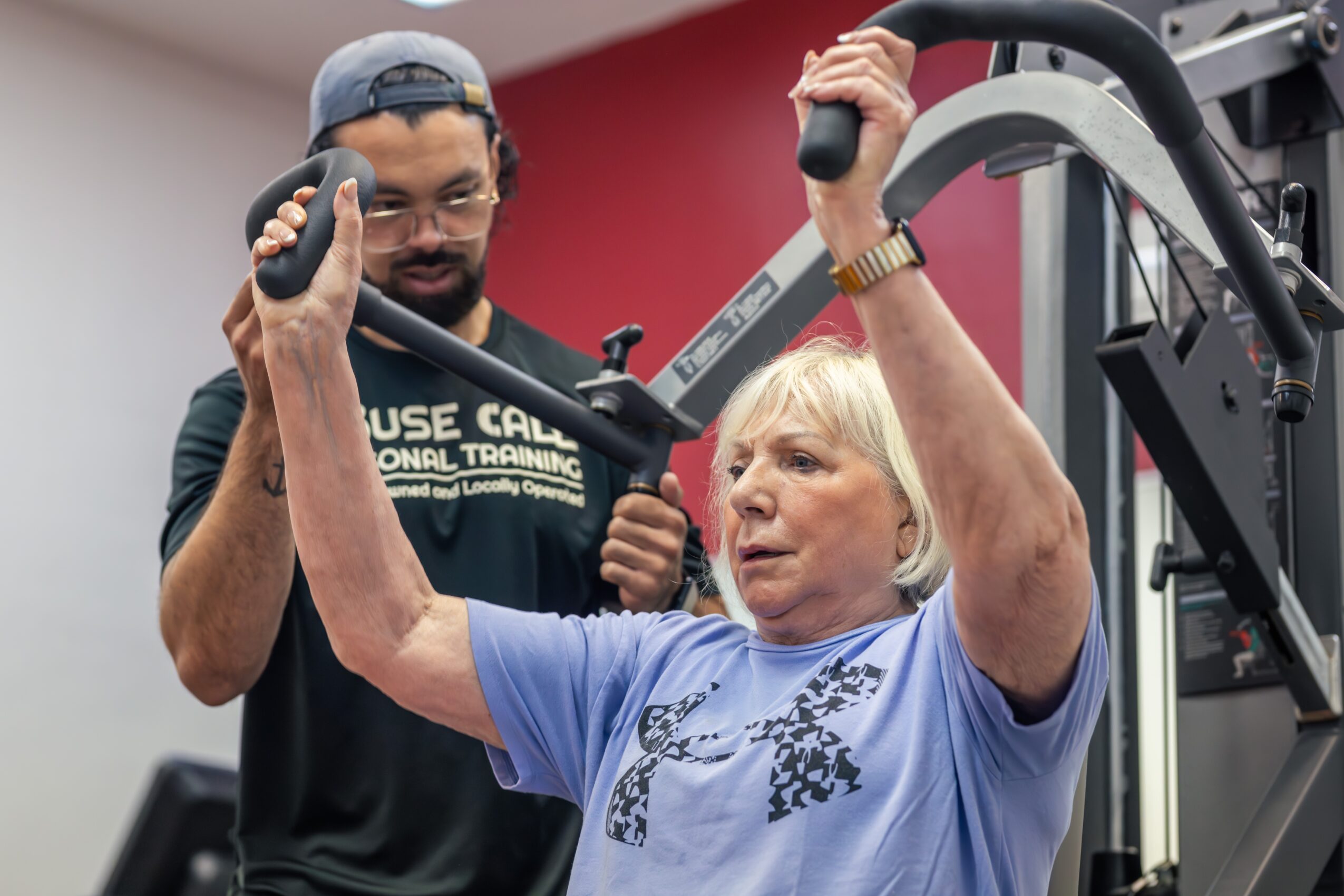 Older woman using a chest press machine with a trainer standing behind her, in a gym setting.