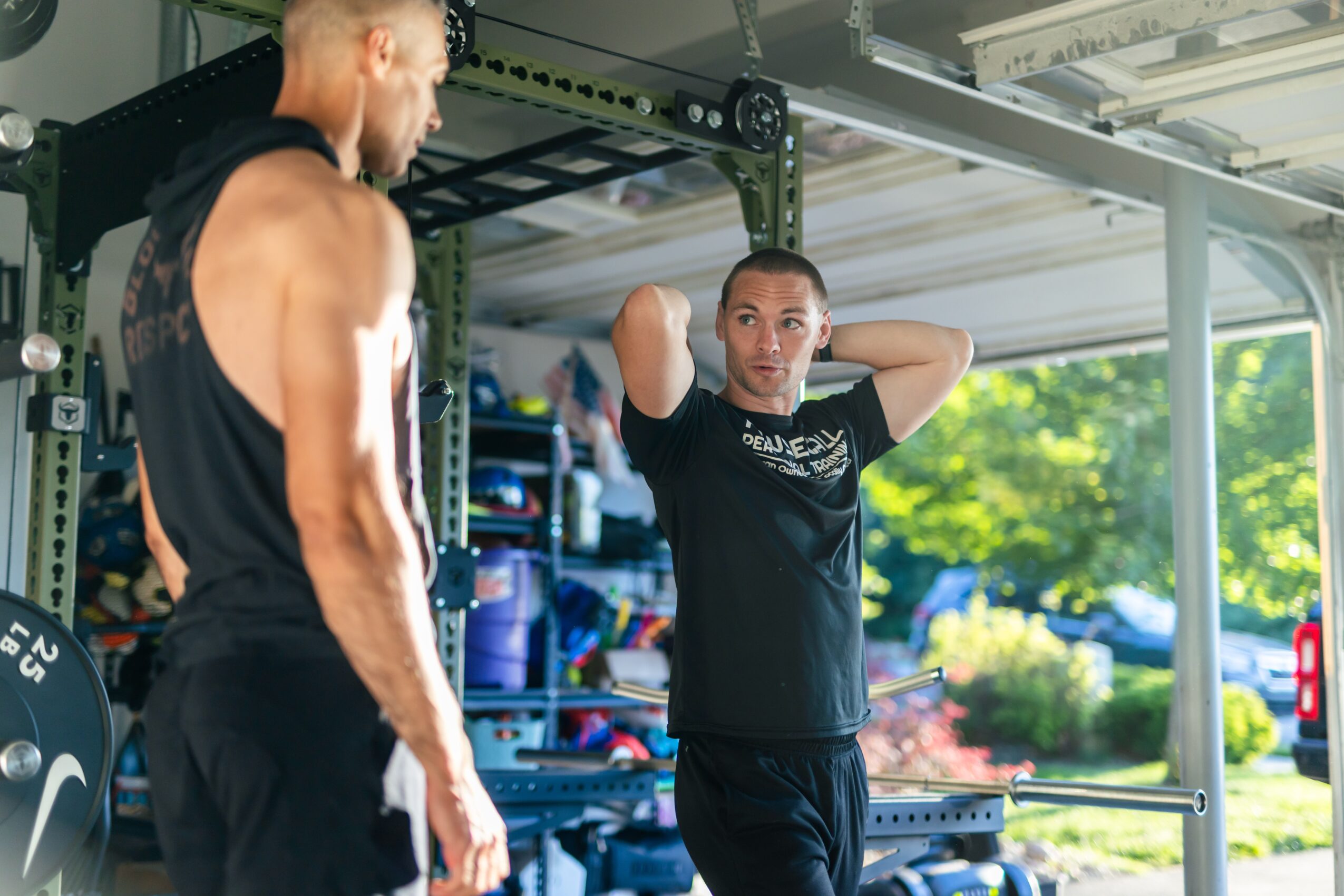 Two men in a gym, one lifting weights and the other resting with hands behind head, with gym equipment and outdoor view.