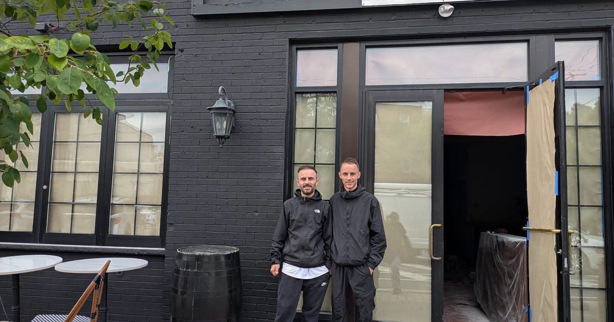 Two men stand outside a black building with large windows and an open door, with a trash bin nearby.
