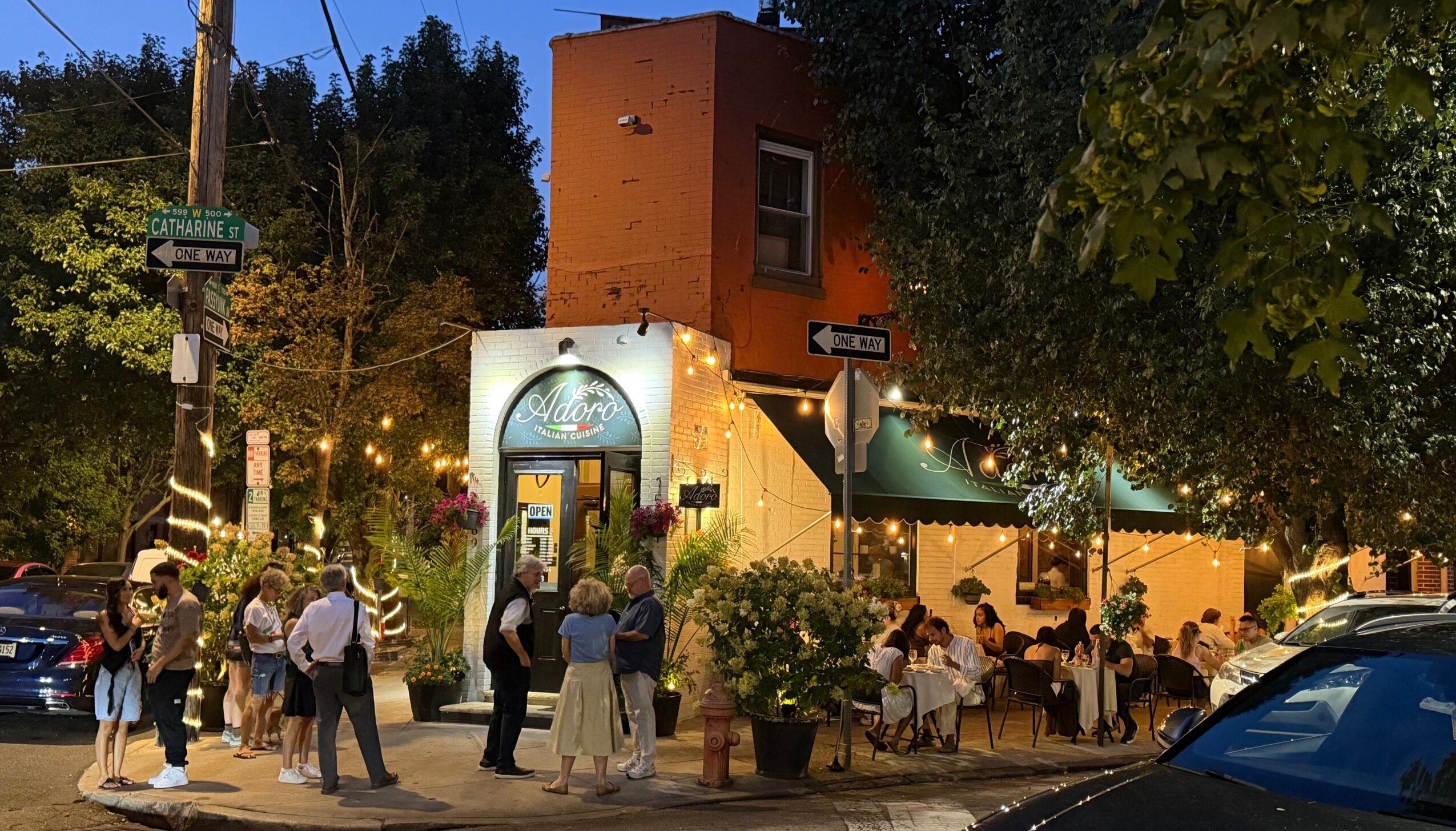 People gather outside a building with string lights and outdoor seating, trees and parked cars nearby, evening setting.