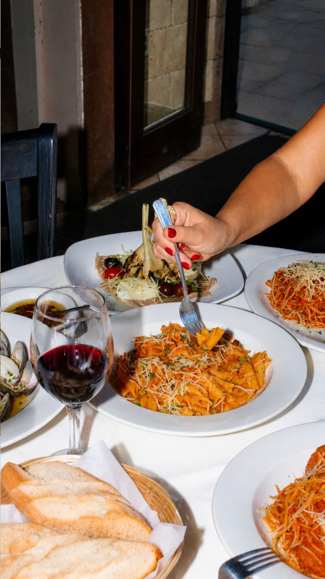 Person's hand holding fork over plate of spaghetti, with other plates of pasta and a glass of red wine on table.