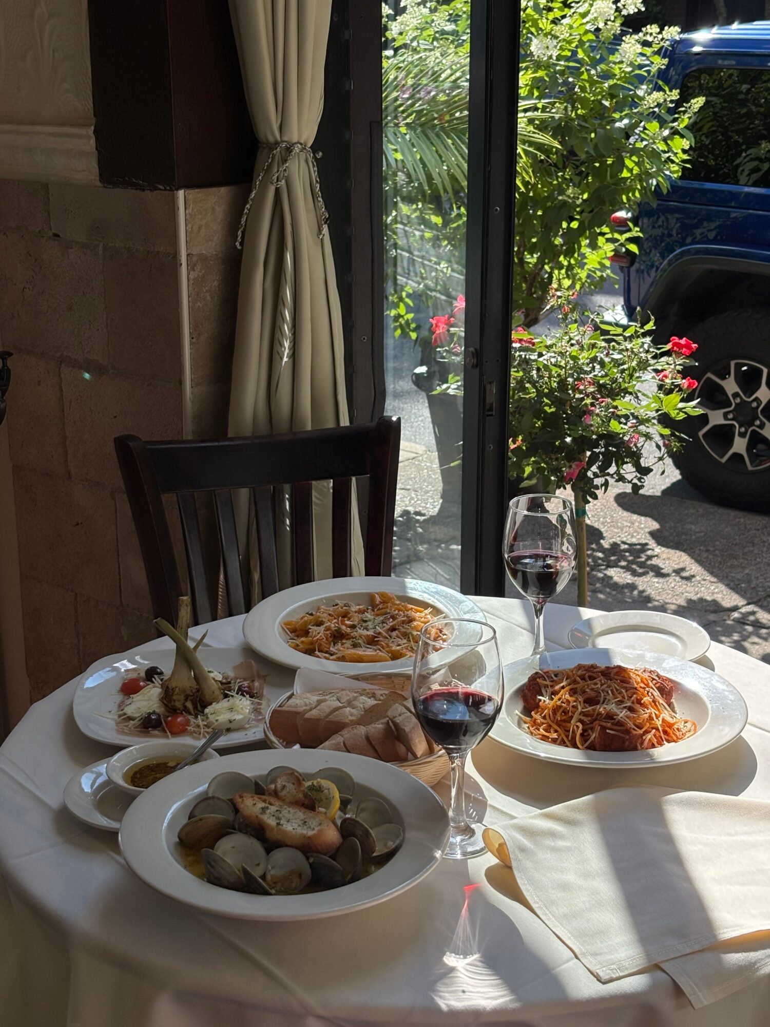 Table set with pasta dishes, wine glasses, and bread near a window with outdoor greenery.