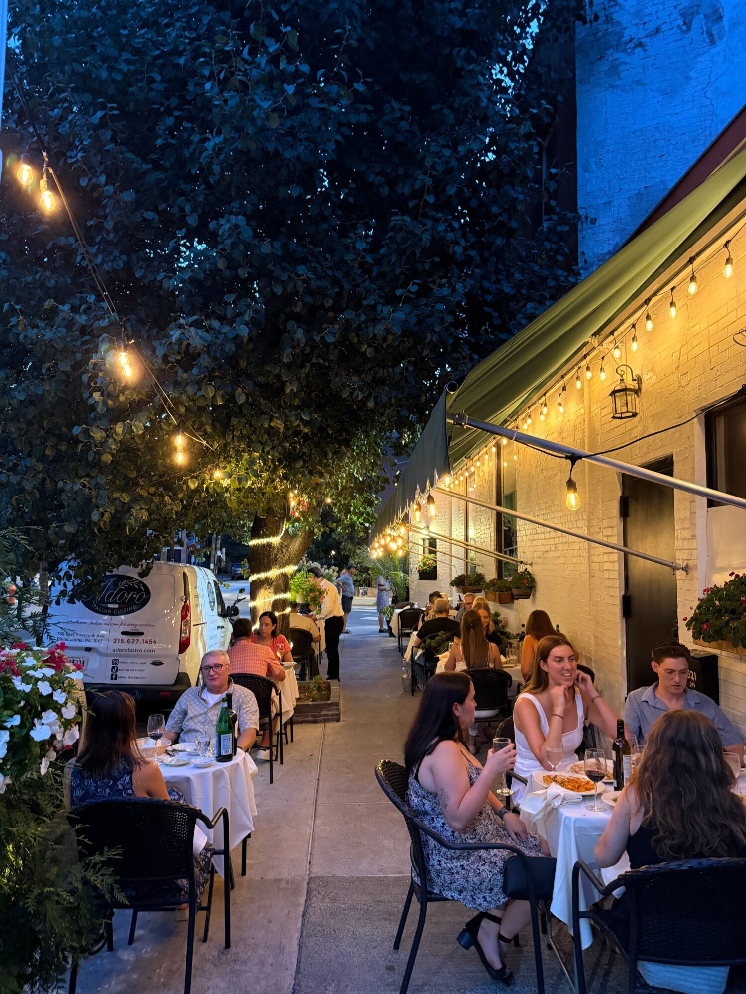 People sitting at tables outdoors during evening, with trees and string lights in background.