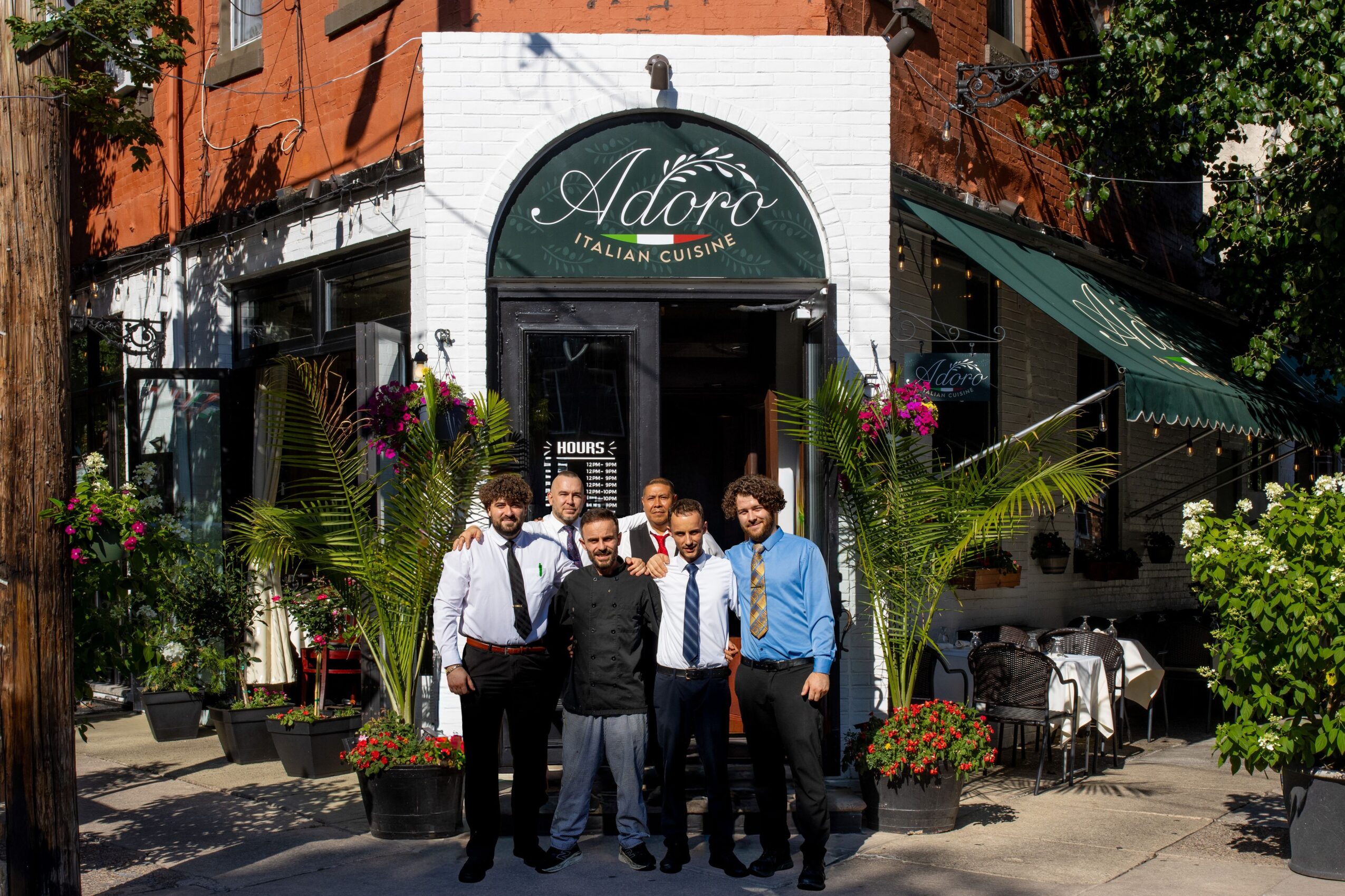 Group of six people standing outside a restaurant with greenery and flowers, smiling, in front of a sign that says 'Adora Italian Cuisine'.