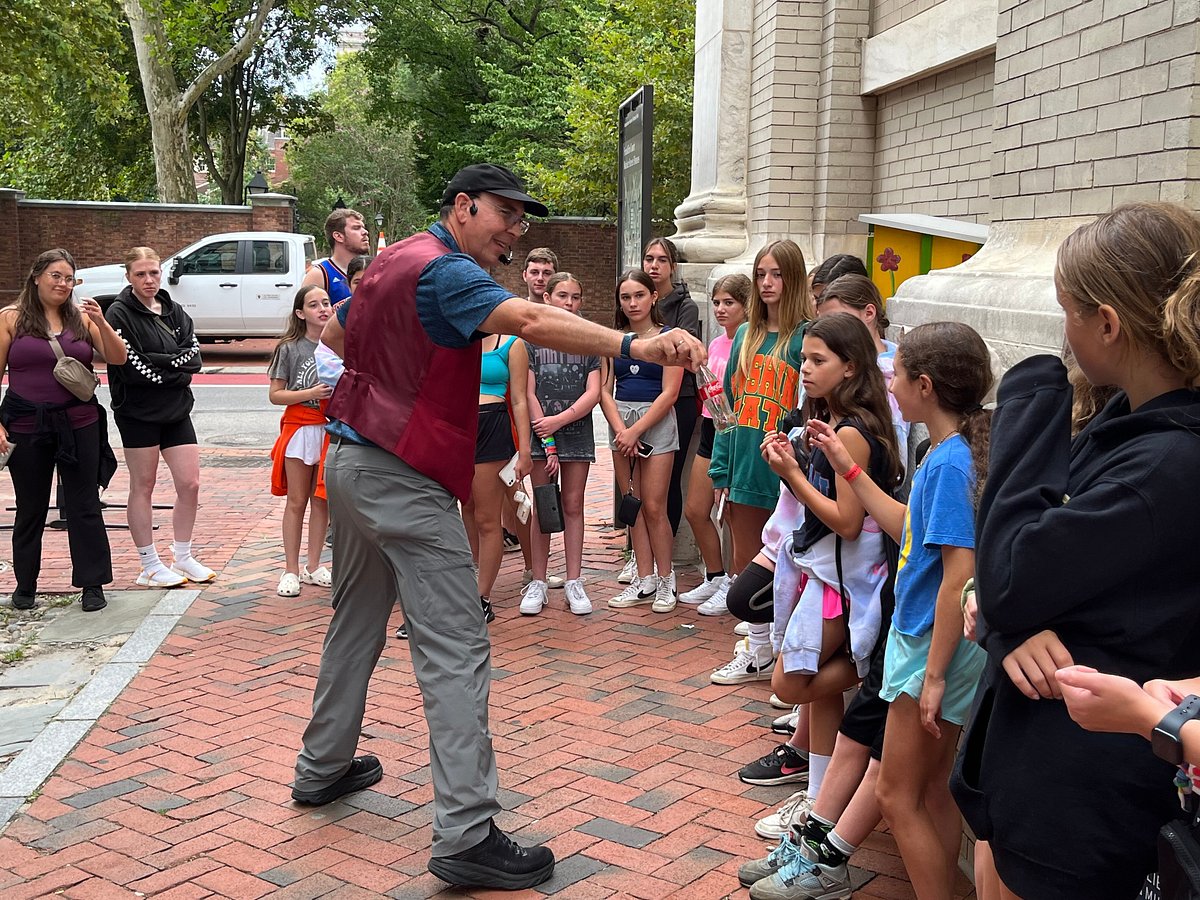 A man performs a magic trick for a group of children outdoors on a brick sidewalk, with trees and a building in the background.