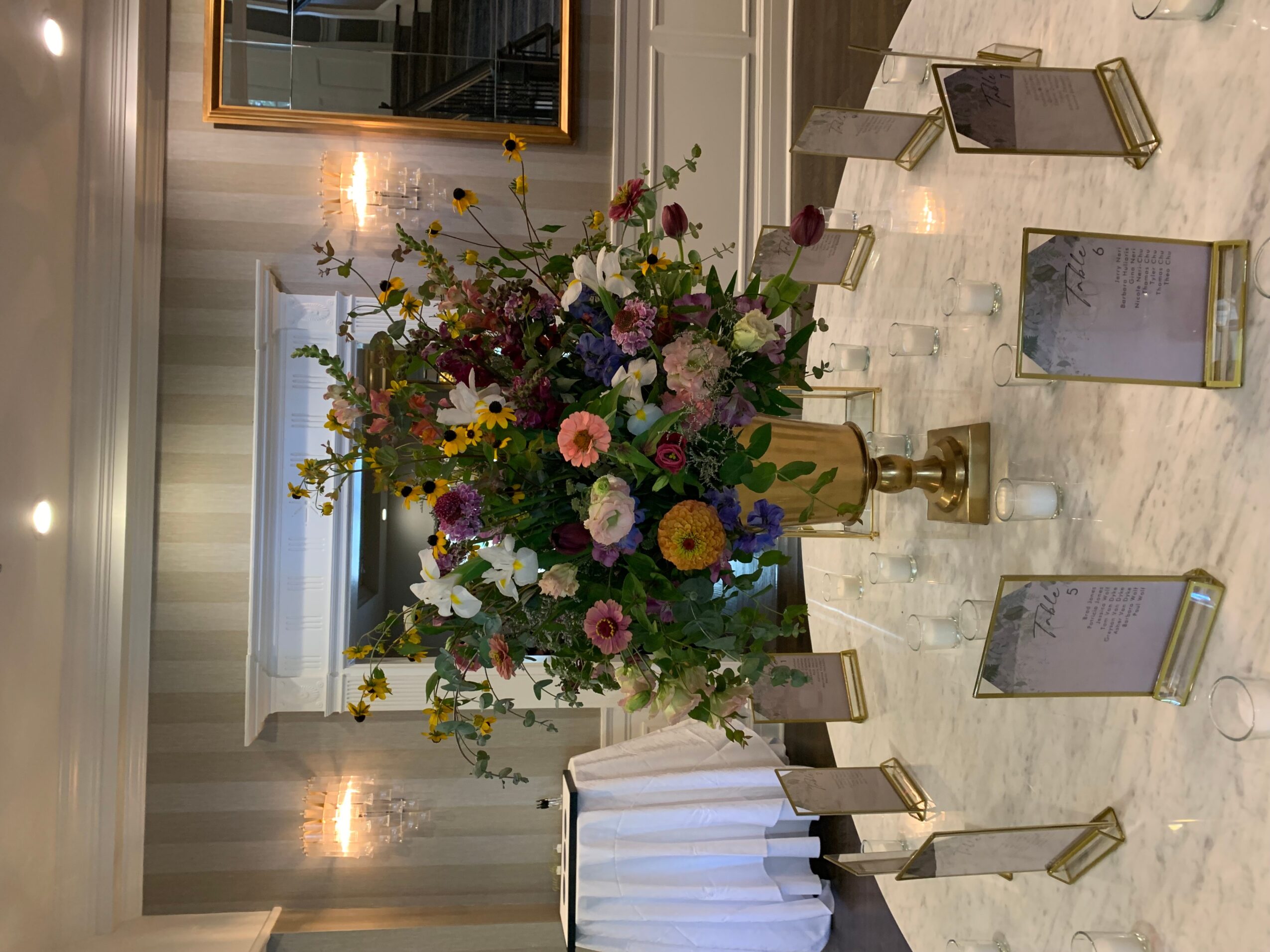 Large colorful flower arrangement on a marble table with small framed photos, in a well-lit room with curtains and a mirror.