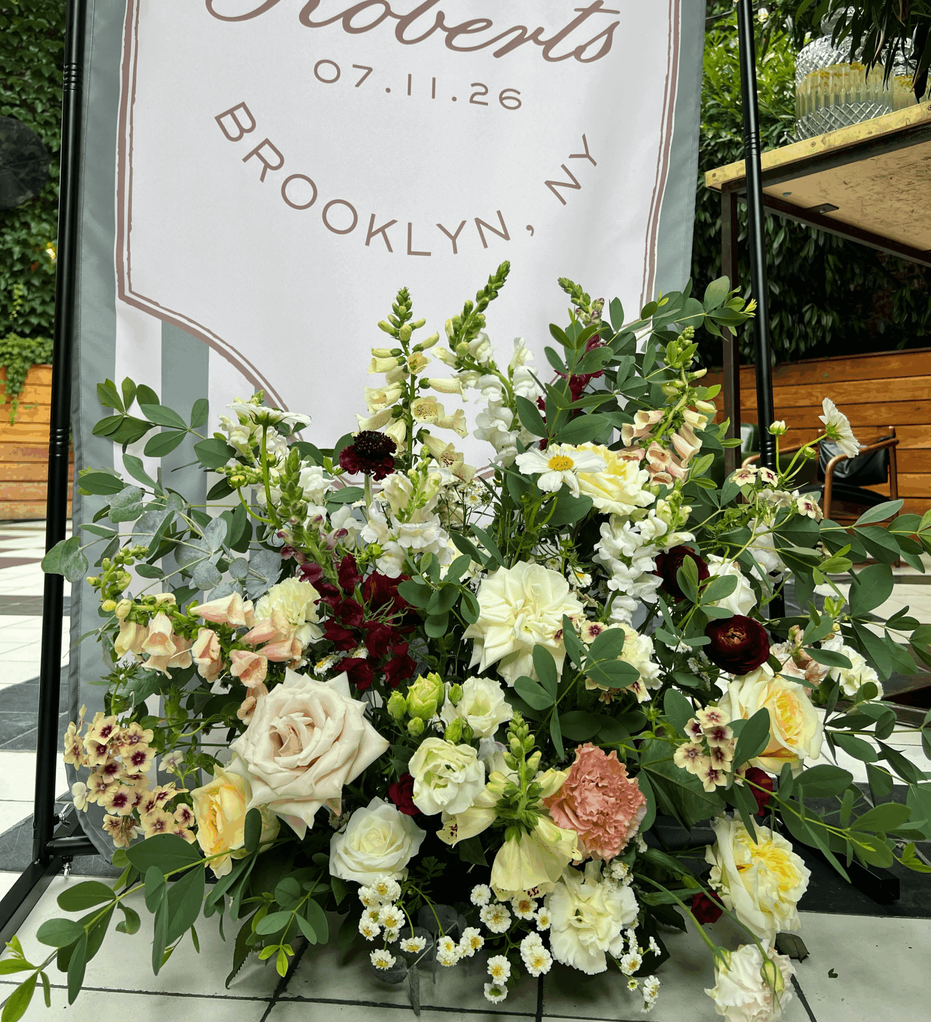 Flower arrangement with roses, greenery, and other flowers in front of a sign with text and date.