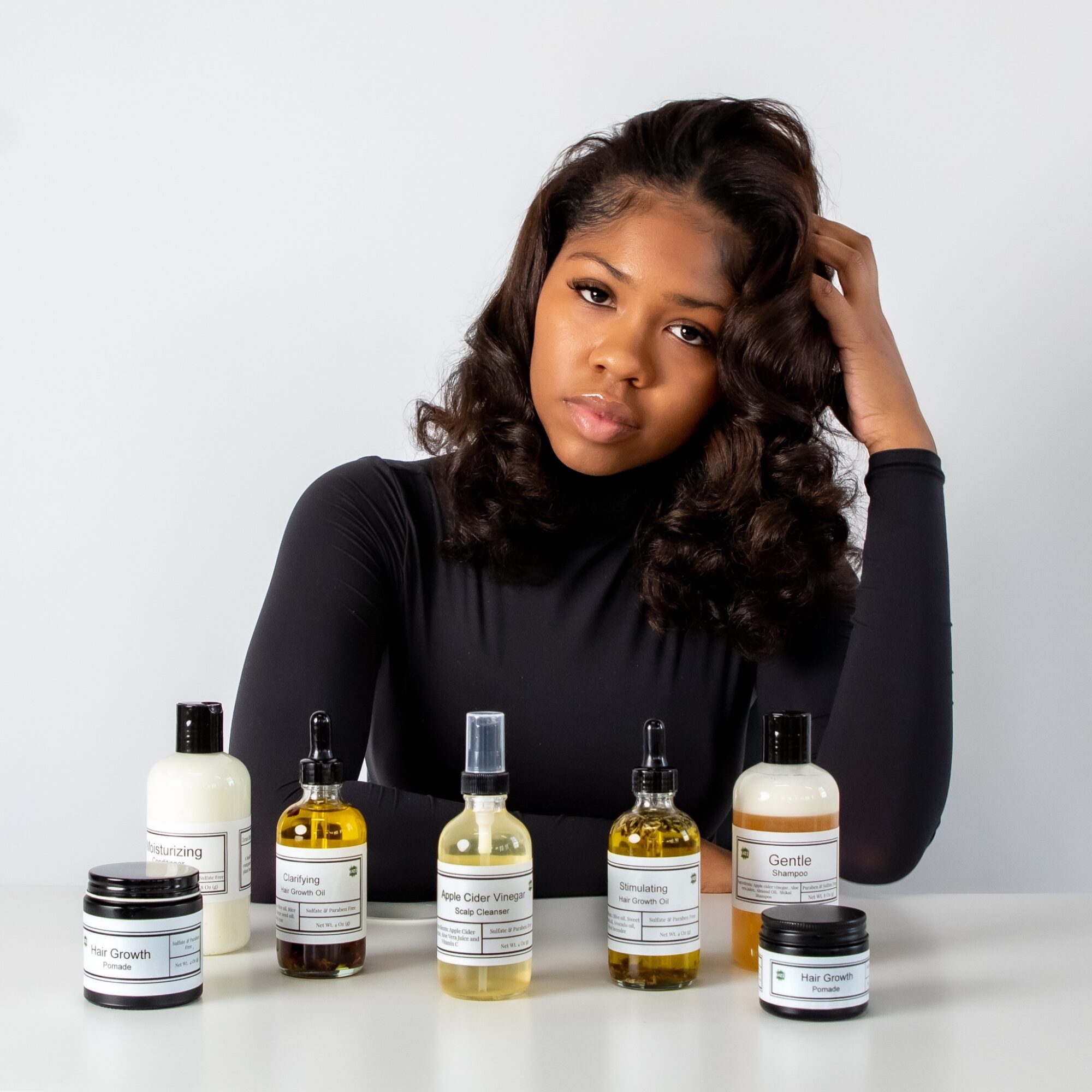Young woman with wavy hair in black top, sitting at table with skincare products, looking at camera.