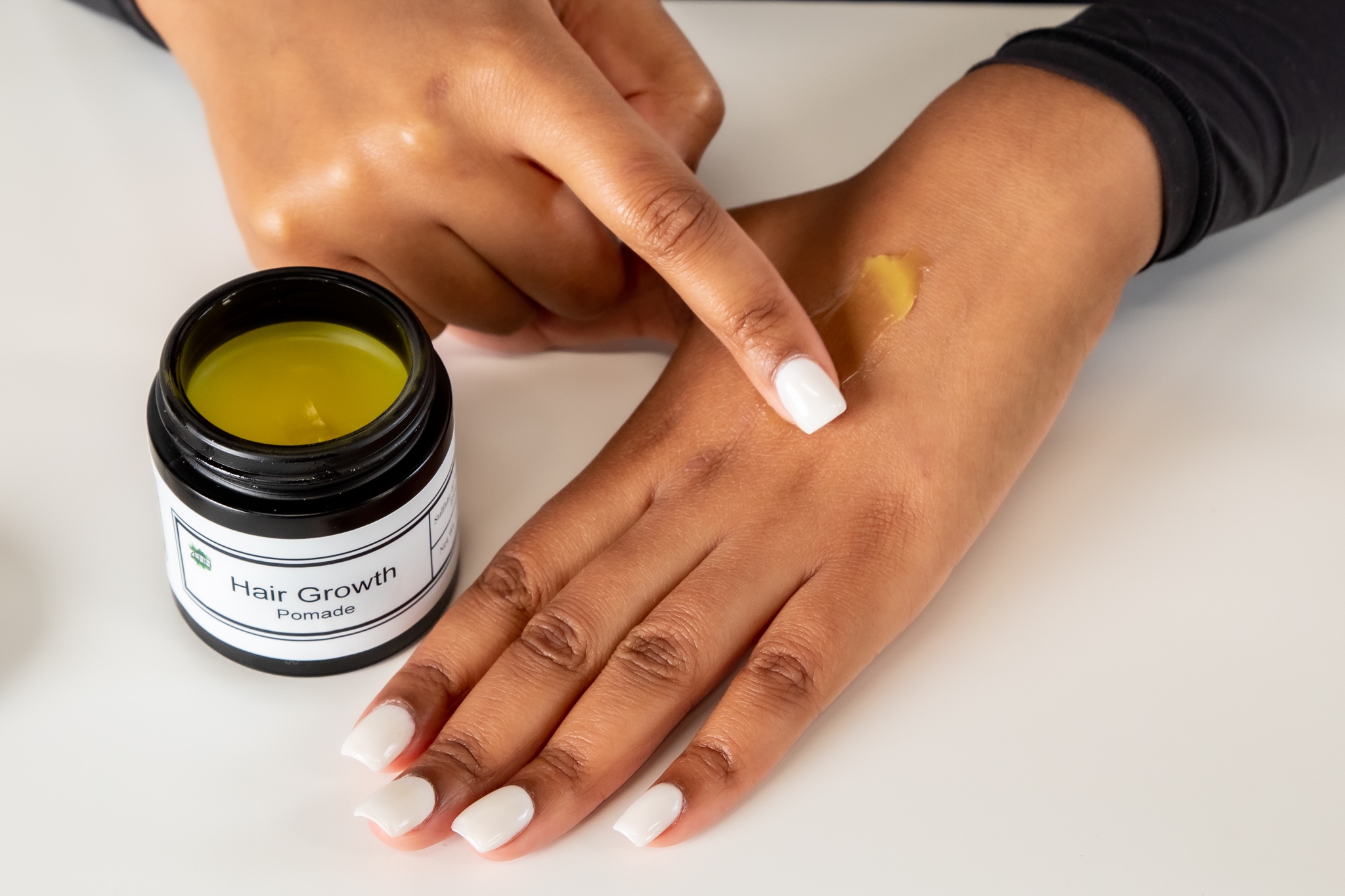 Person applying yellow hair growth product to hand with a finger, next to a jar labeled 'Hair Growth'.