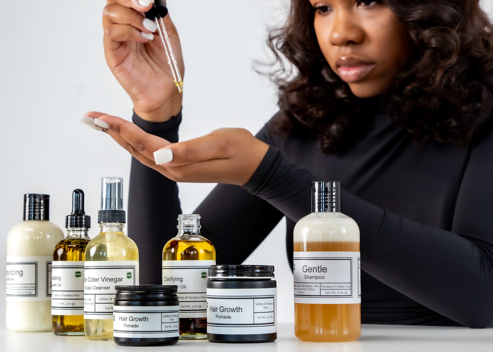 Woman with curly hair holding a dropper over skincare products on a white surface.