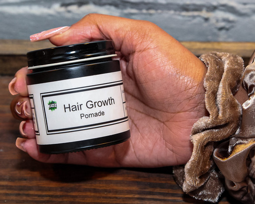 Hand holding a small jar labeled 'Hair Growth Pomade' with a black lid, against a background of water and wood.