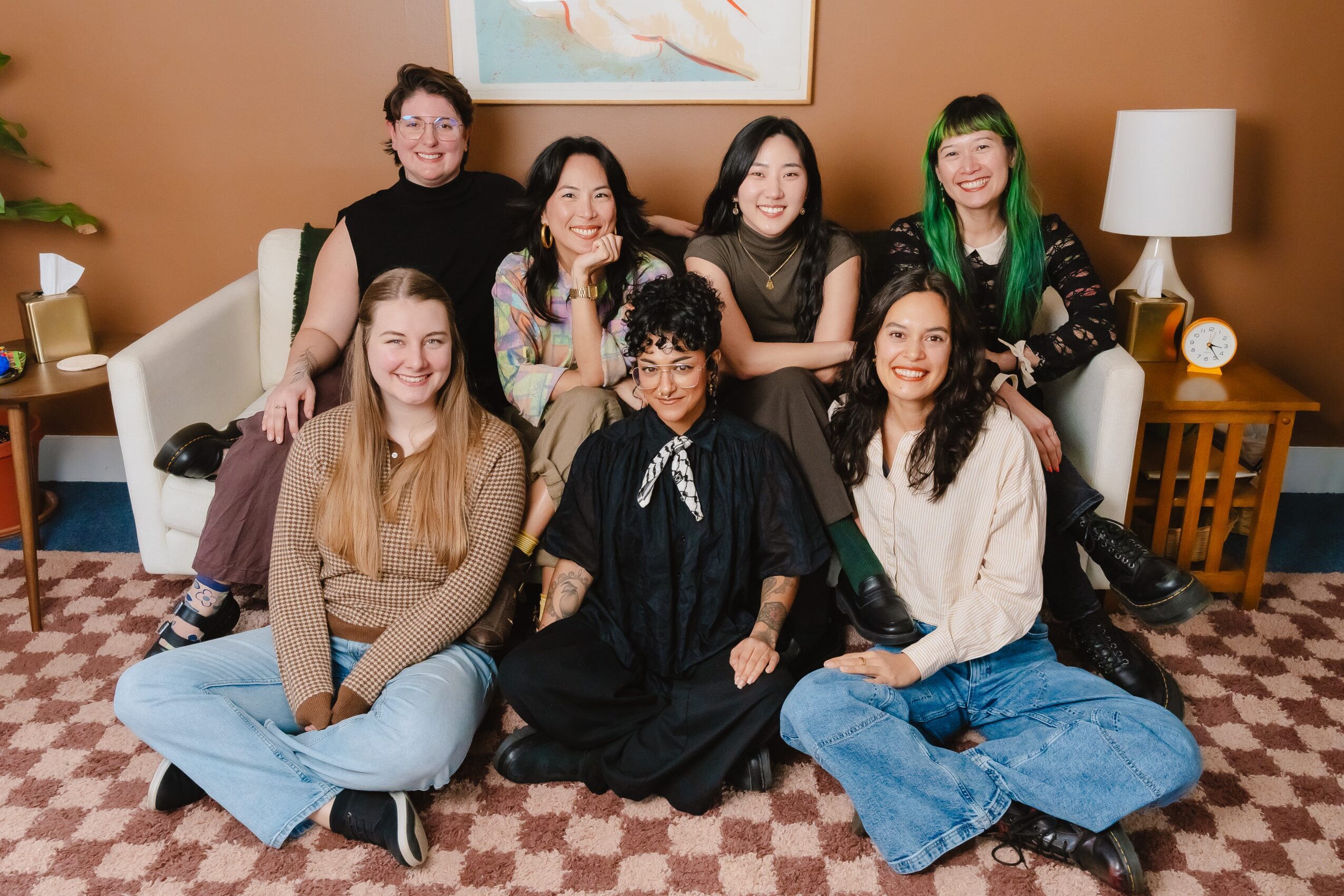 Seven women sitting and standing on a sofa in a living room, smiling, with a painting and lamp in the background.