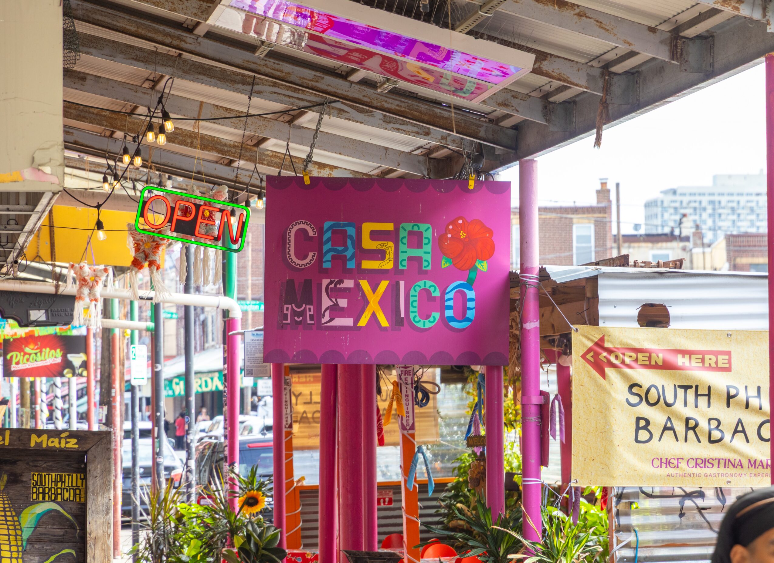 Colorful sign with text 'Casa Mexico' and a red apple, under a covered outdoor area with city buildings in background.