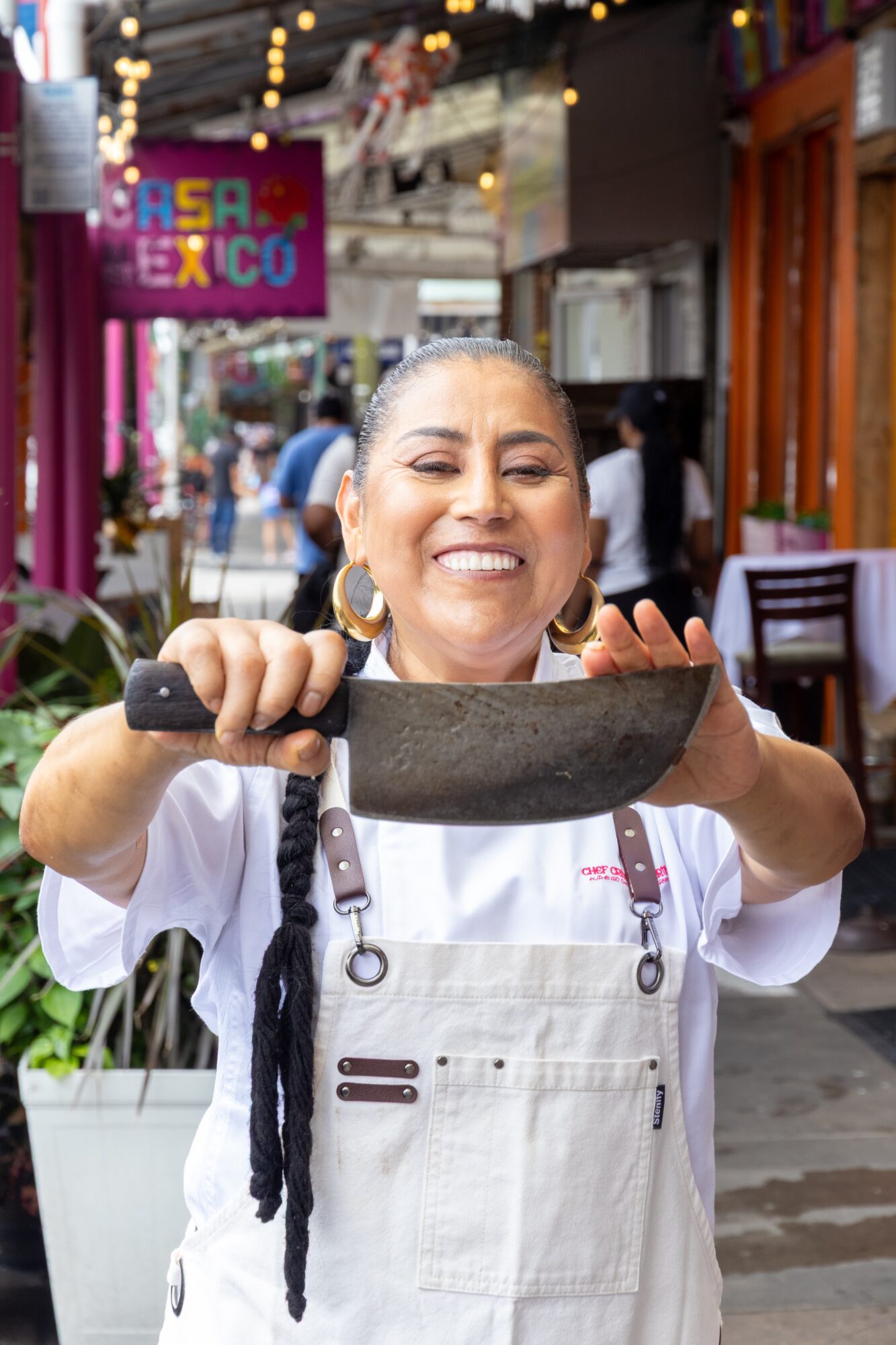 Smiling woman holding a large knife in a lively outdoor market or street scene, wearing a white apron and earrings.