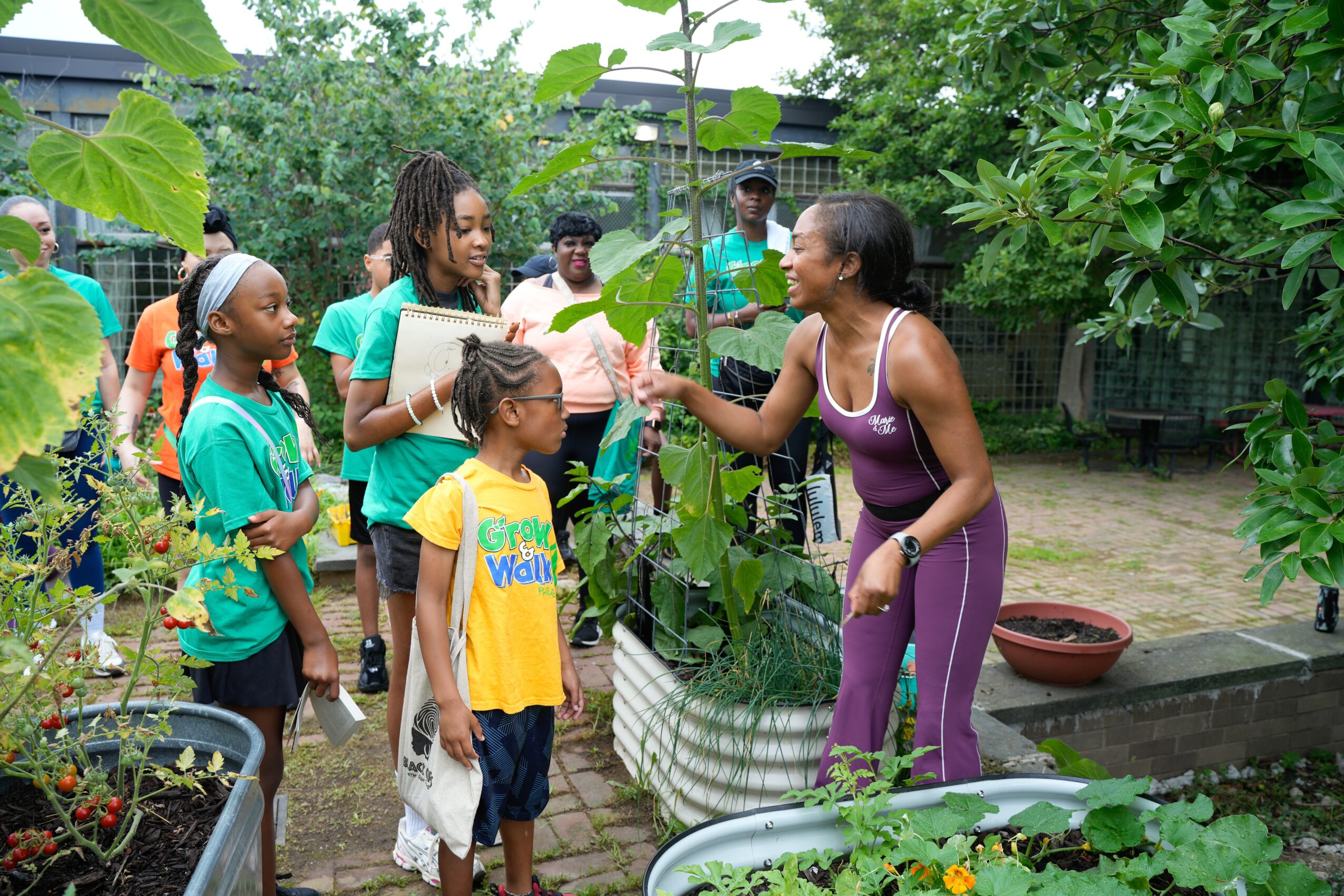 Group of children and an adult woman in a garden, woman talking to a girl, surrounded by plants and trees.