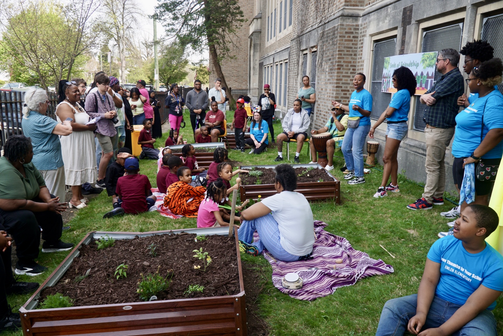 Group of people gathered outdoors around garden beds, some sitting and some standing, with children present, near a building.