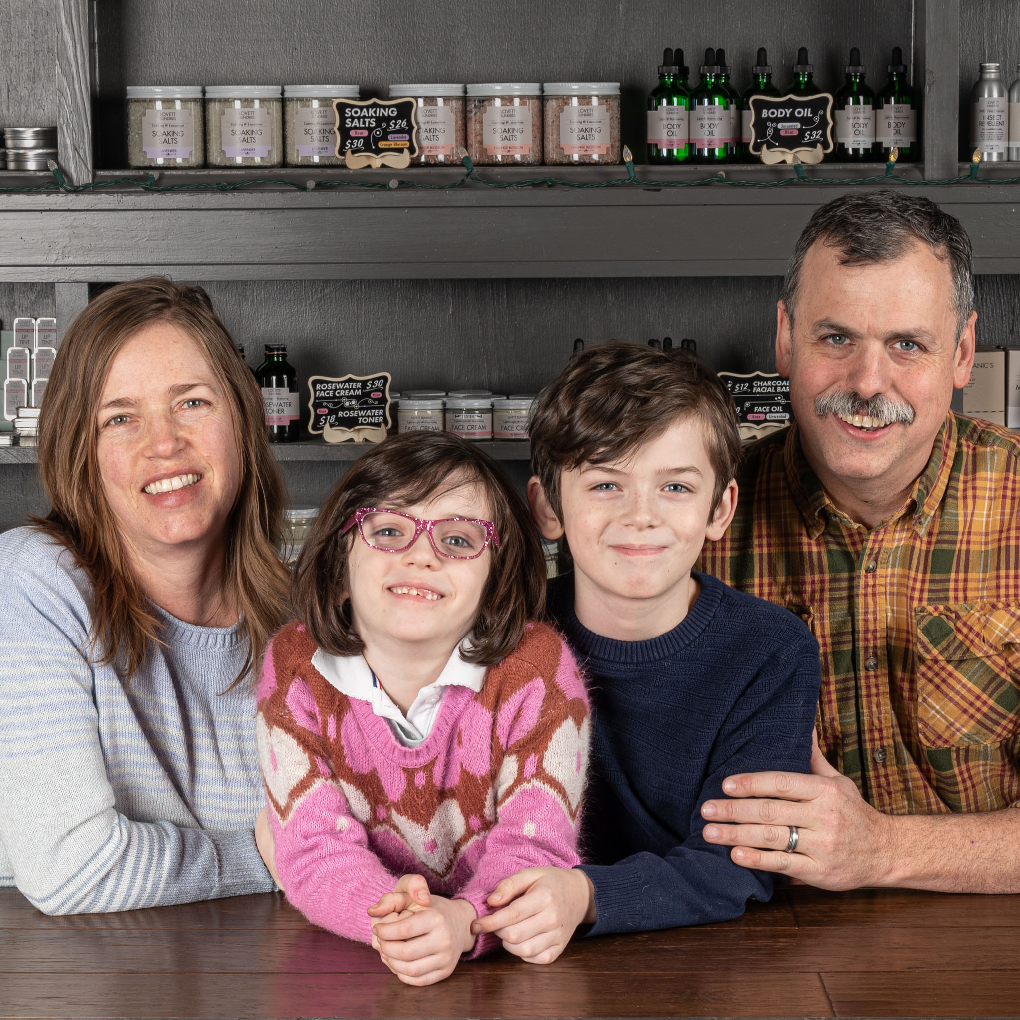 Family of four smiling at a counter in a shop, shelves with products behind them.