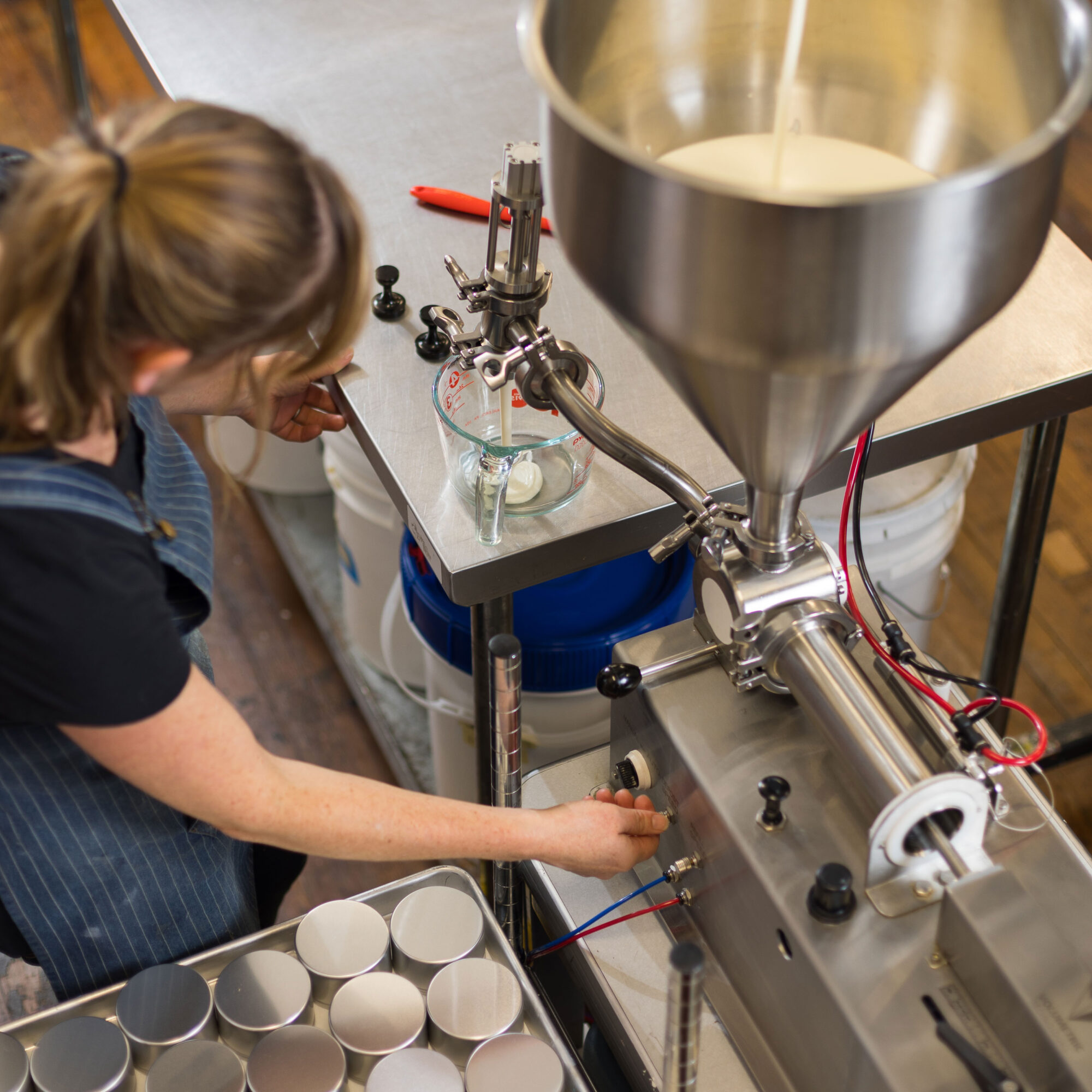 Person operating a machine with a large funnel, pouring liquid into small cups on a table, in a laboratory or workshop setting.