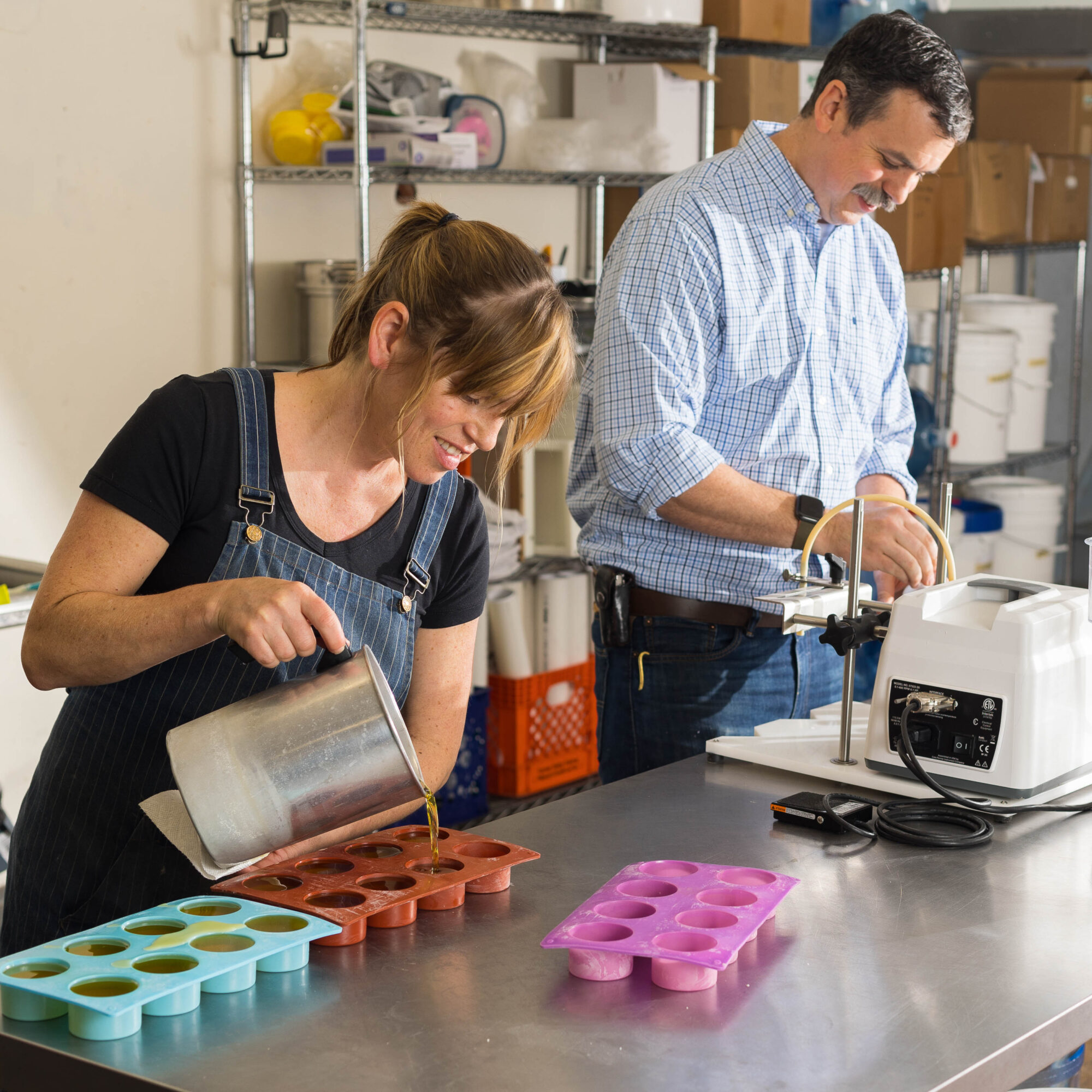 Two people working in a kitchen, pouring liquid into silicone molds on a stainless steel counter.