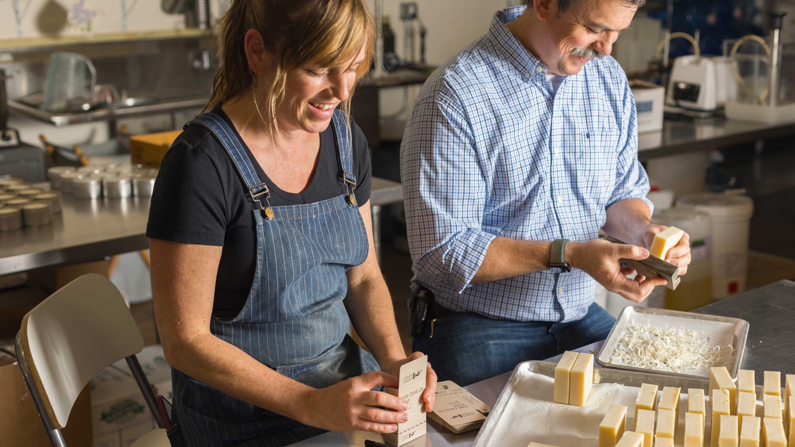Two people working with soap bars in a kitchen or workshop, smiling and handling soap blocks and molds.