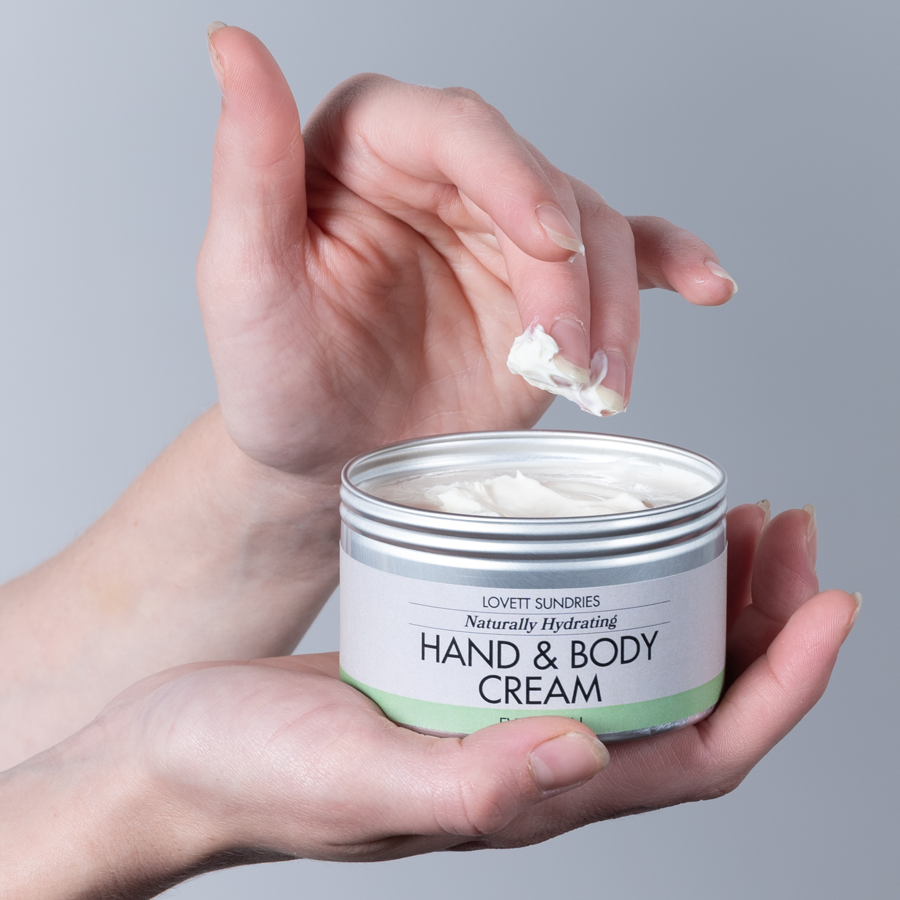 Person holding a jar of hand and body cream, with a finger applying cream, against a plain background.