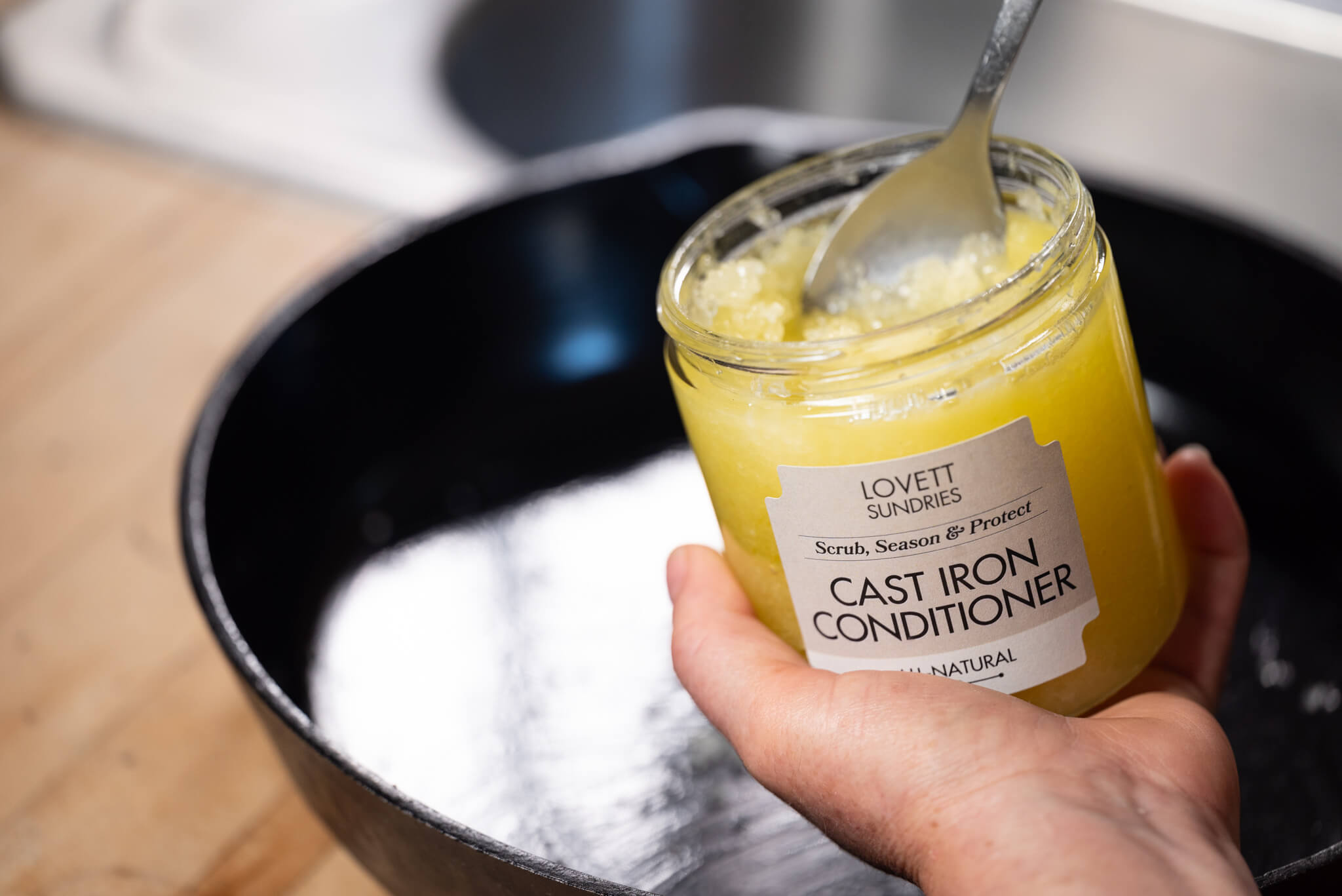 Hand holding a jar of cast iron conditioner over a black skillet, with a spoon inside the jar, on a wooden surface.