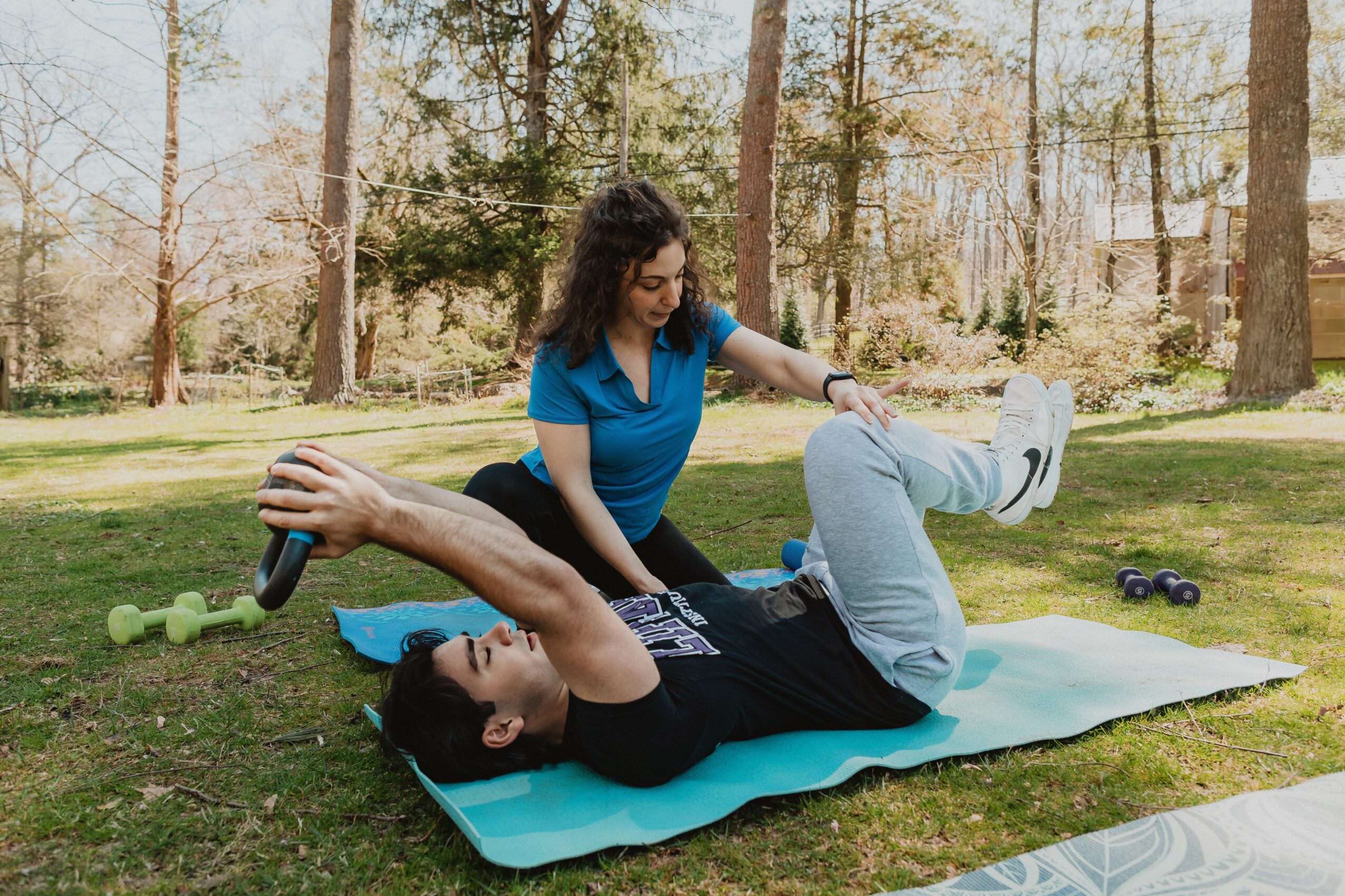 Woman assisting man with exercise on a mat outdoors, surrounded by trees, with dumbbells nearby.