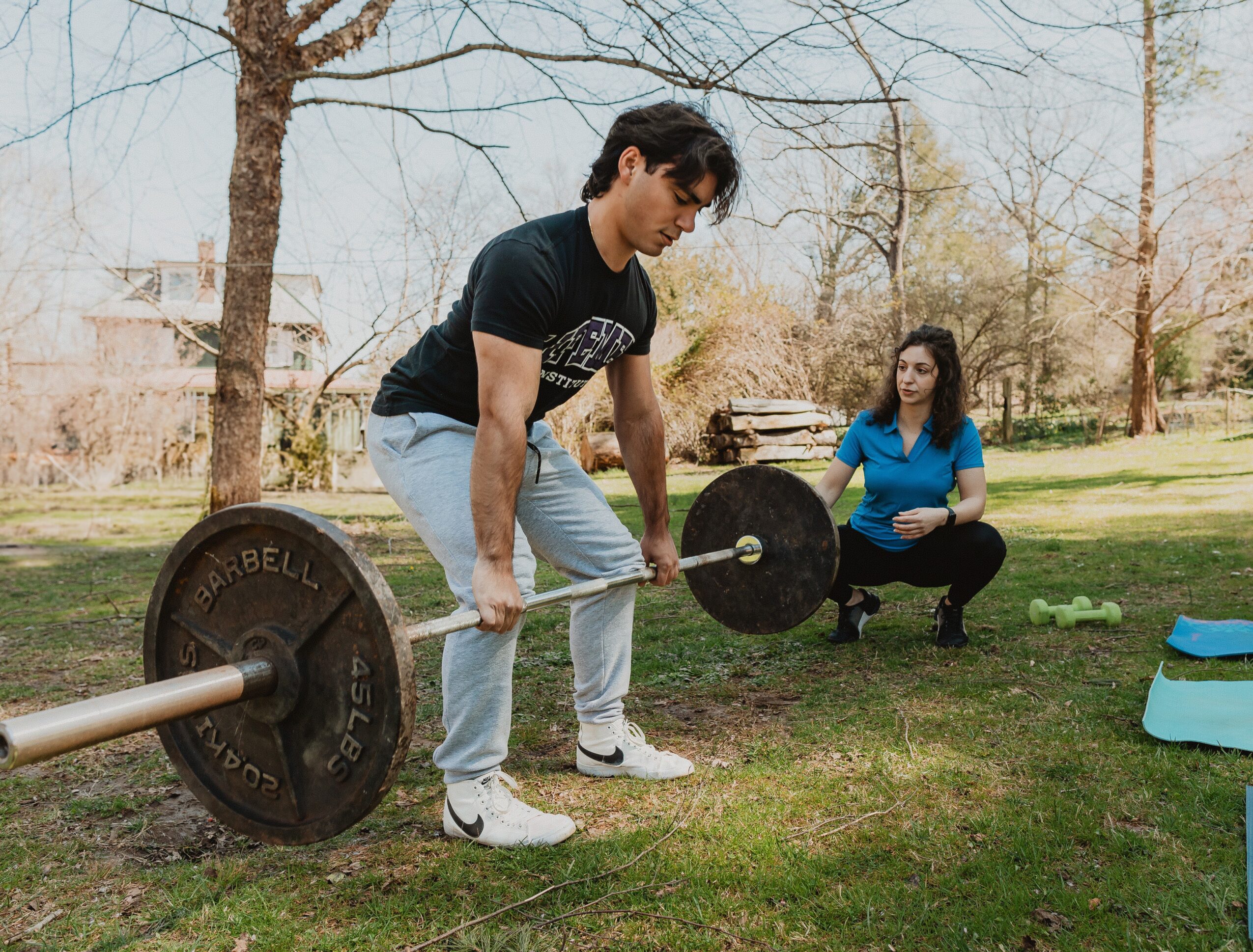 Man lifting a barbell outdoors with a woman sitting on the grass nearby, trees in the background.