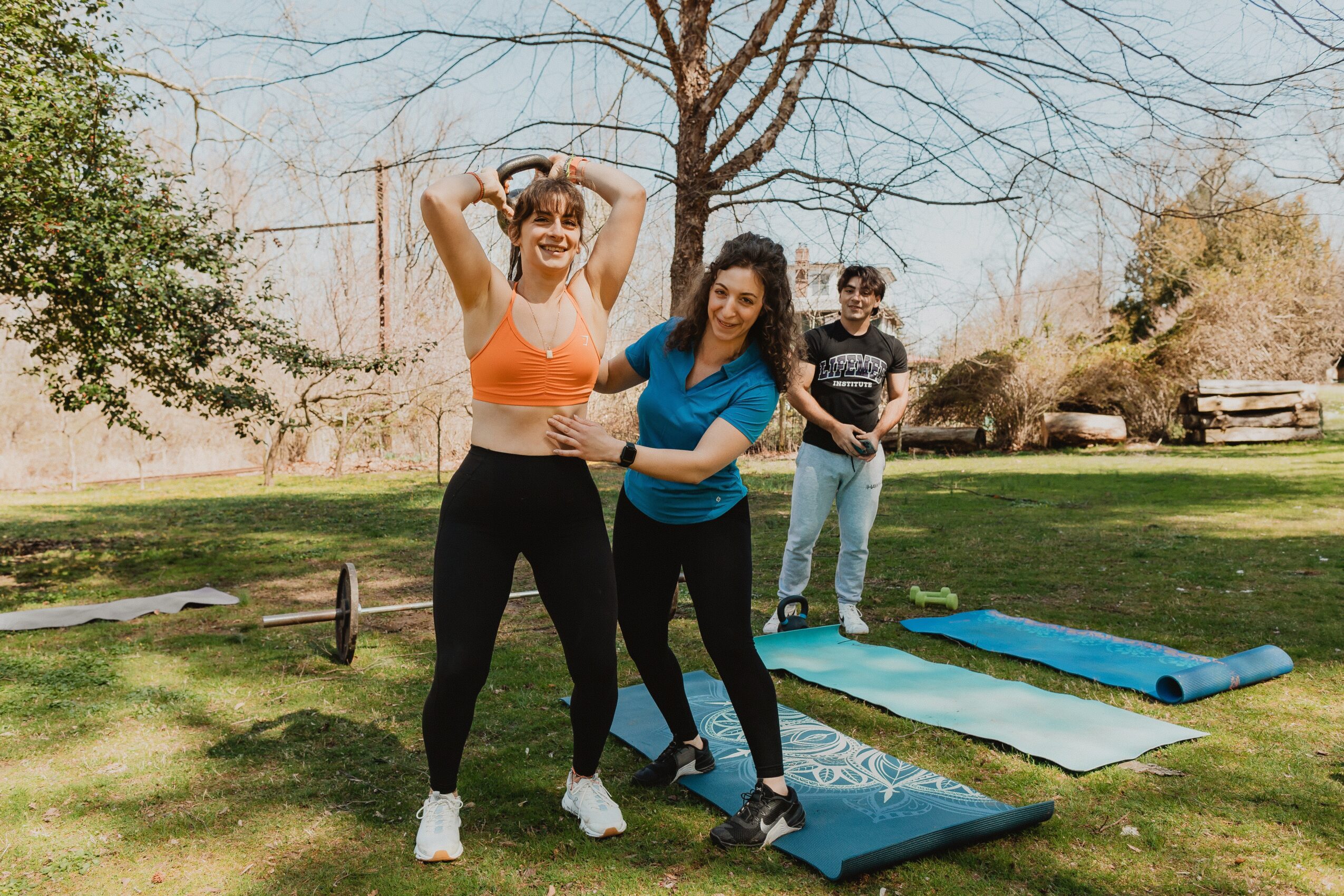 Three women outdoors on grass with yoga mats, trees, and benches in background, smiling and posing.